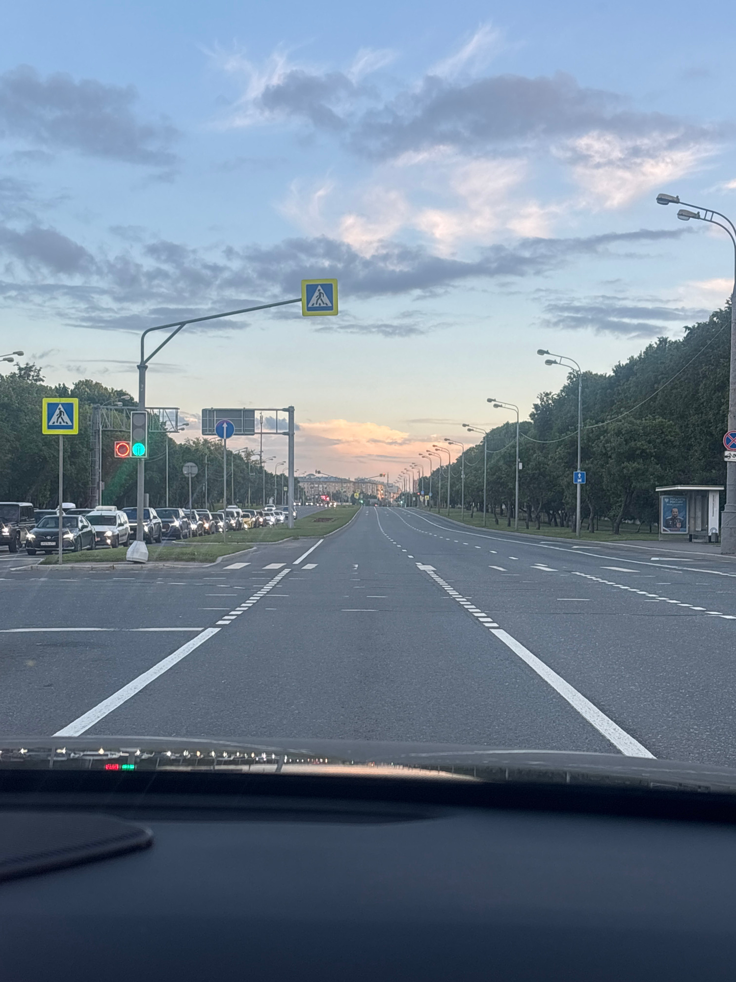 This is a medium-distance, eye-level, eye-level, street-level photograph taken from inside a vehicle, looking forward. The scene captures a multi-lane road lined with trees and streetlights under a cloudy sky at dusk. On the left side of the road, there is a traffic light showing a green signal and a pedestrian crossing sign. A queue of cars is visible on the left lane. On the right side of the road, there are more streetlights and a bus stop. The road is empty in the distance, suggesting it leads to the city center. The overall atmosphere is calm and serene, with the soft light of dusk creating a peaceful ambiance. The composition is balanced, with the road leading the viewer's eye into the distance. The colors are muted and harmonious, with the blue sky, green trees, and gray asphalt creating a natural palette. The image could be taken at Vernadsky Avenue in Moscow, Russia, given the wide avenues and presence of trees and streetlights.