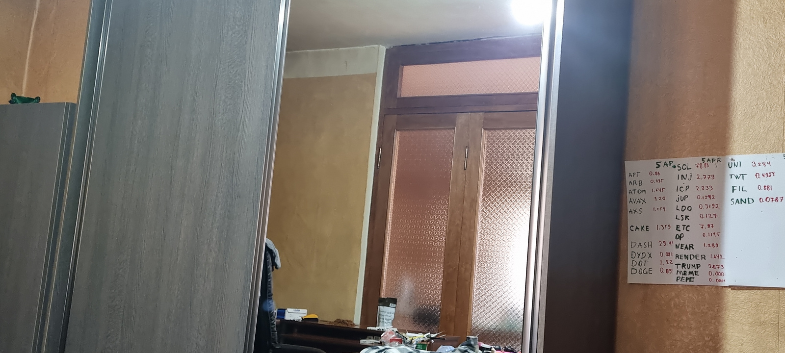 This is a medium shot, interior photograph taken in Tbilisi, Georgia. The scene depicts a room with a large, light-grey, wooden wardrobe or closet dominating the left side of the frame. The wardrobe has vertical paneling and polished metal trim. To the right of the wardrobe, a doorway with a dark wood frame and textured glass panels is visible, leading to another room. The wall behind the wardrobe and to the left of the door is painted a warm, sandy yellow.
On the right side of the frame, a bulletin board or whiteboard is affixed to a textured, light brown wall. This board contains handwritten lists of what appear to be cryptocurrency tickers and their corresponding prices, dated "5 APR". Examples of visible text include "APT 0.86", "ATOM 1.645", "SOL 78.83", "UNI 3284", "ICP 2.233", and "SAND 0.0787".
In the foreground, near the bottom of the image, several objects are scattered on what might be a shelf or surface in front of the doorway. These include items that resemble packaging, a small yellow object, and parts of other items that are out of focus. A small, green toy airplane is resting on the top edge of the wardrobe on the far left. The lighting in the room appears to be artificial and somewhat dim, with a bright light source visible at the top of the frame, possibly from a ceiling fixture reflecting off the wardrobe. The overall impression is of a lived-in space, perhaps a bedroom or living area, with elements of personal organization or tracking evident on the whiteboard. There are no people visible in the image.