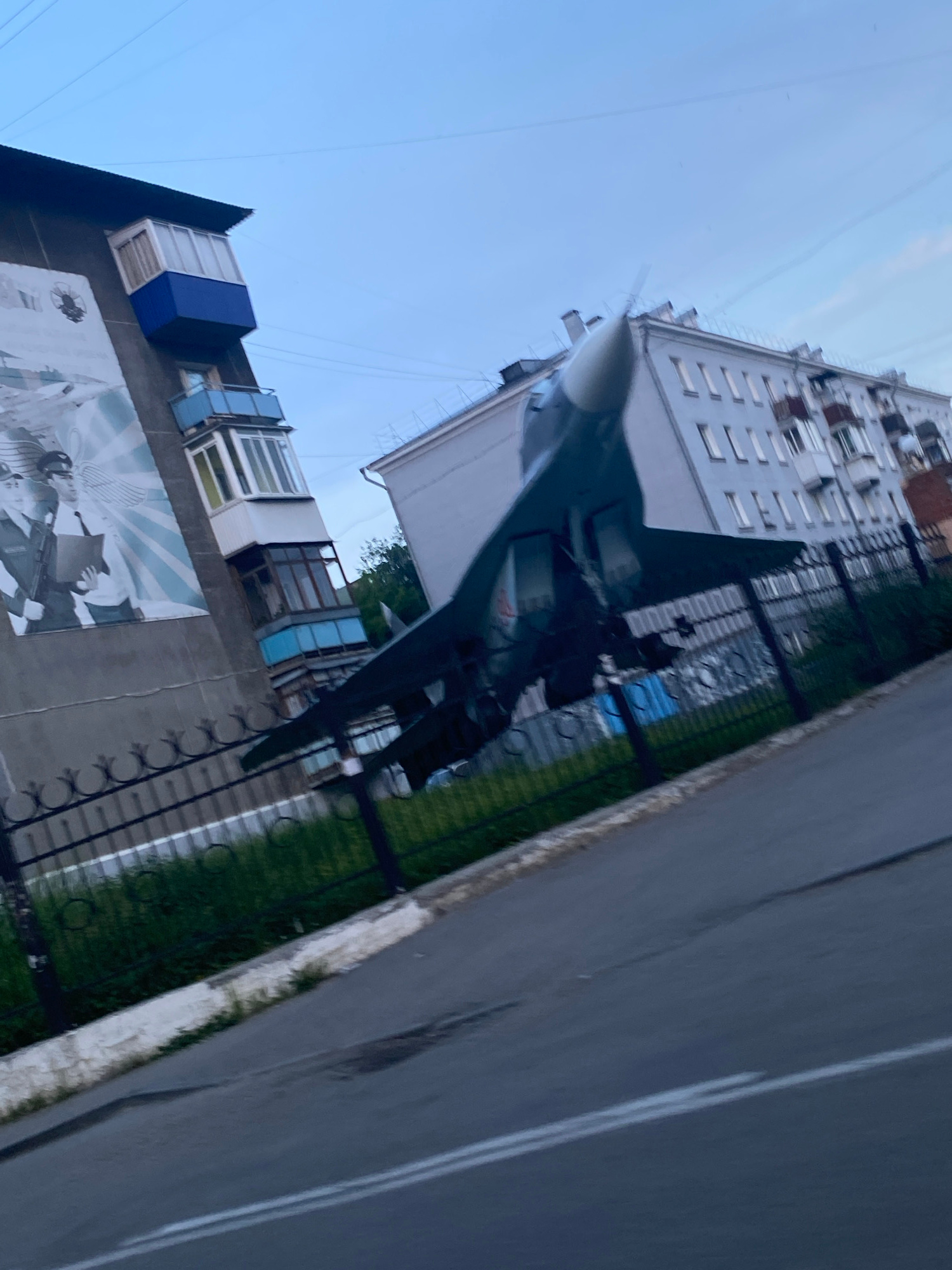 The image depicts an outdoor scene, likely in the city of Irkutsk, Russia, given the context provided. The primary subject is a dark-colored fighter jet, positioned at an upward angle, with its nose cone pointing towards the right and slightly upwards. The jet is situated in front of a light-colored apartment building with multiple windows and balconies. To the left of the jet and apartment building is a gray building adorned with a large mural. The mural appears to be a black and white depiction of two individuals in uniform, possibly military or law enforcement, with wings behind them. A black metal fence with decorative elements runs horizontally across the middle ground of the image, separating the buildings from a grassy area. A paved road with white markings occupies the foreground, sloping upwards from the bottom right corner of the frame. The sky above is a pale blue, suggesting it might be early morning or late evening. The overall impression is of an urban environment with a monument or display featuring a military aircraft.