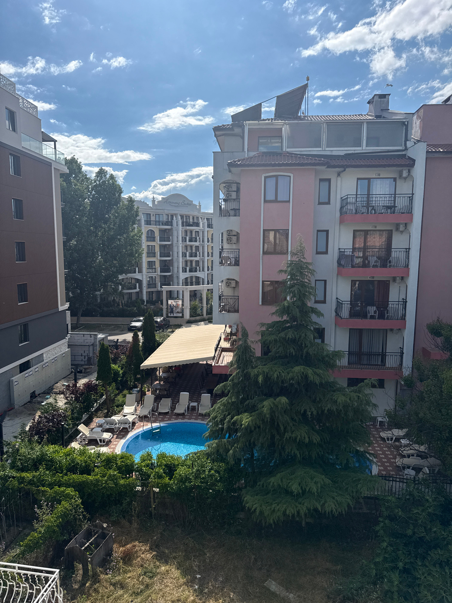 The image, taken in Nesebar, Bulgaria, depicts an outdoor scene with a swimming pool and surrounding buildings. The foreground features a small, blue swimming pool with lounge chairs arranged around it. Lush green bushes and trees border the pool area. In the background, there are several multi-story buildings. One building is painted pink and white with balconies and dark railings. Another taller building, with a light yellow facade and white decorative elements, is visible further in the background. The sky is blue with scattered white clouds, suggesting a sunny day. The overall impression is that of a resort or residential complex.