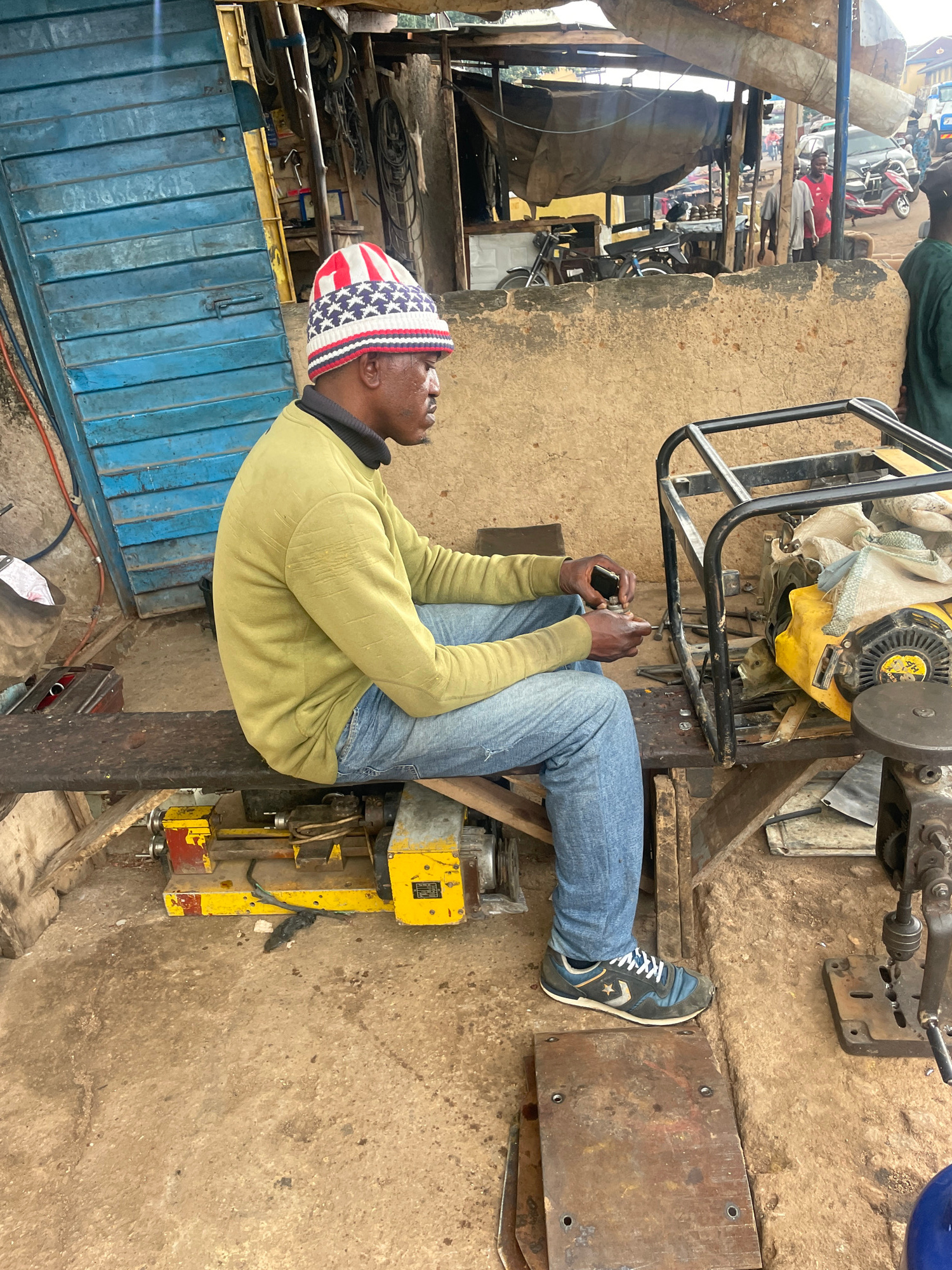 The image shows a person sitting in an outdoor workshop in Jos, Nigeria. The person is wearing a green long-sleeved shirt, blue jeans, a knit cap with a red, white, and blue star pattern, and sneakers. They are holding a small black object in their hands and appear to be working on or examining a yellow and grey piece of machinery. The machinery is positioned on a wooden platform, and other tools and equipment are visible in the background. The setting appears to be a street-side workshop with a blue metal gate and a rough concrete floor. The lighting suggests it is daytime, and the weather appears to be overcast or shaded. The person's posture and focus indicate concentration on the task at hand. No visible text is present in the image.