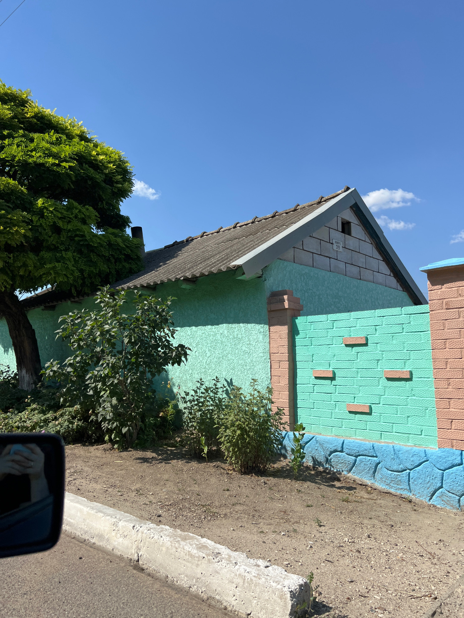 The image shows a residential building with a teal-colored stucco exterior and a gray, corrugated roof. A portion of the roof shows exposed brickwork at the gable end. In front of the building, there is a fence constructed with teal brick on the lower half and light pink brick on the upper half. The teal section of the fence has five horizontal rows of protruding pink brick accents. A light blue decorative band is visible along the very bottom of the teal fence section.
To the left of the building, a large, leafy green tree stands beside the structure, with its trunk partially visible. Various green bushes and plants are growing in front of the building and along the fence line.
In the foreground, a dirty, dry patch of ground is visible, with some scattered small stones and sparse vegetation. A concrete curb runs along the bottom of the frame, separating the dirt area from an asphalt road. A car's side mirror is reflected in the lower-left corner of the image, showing a person's hands holding a light blue object.
The scene is illuminated by bright sunlight, casting shadows on the ground and the building. The sky is clear blue with a few small white clouds. The overall impression is of a sunny day in a residential area. The location is identified as Tashlyk, Moldova.