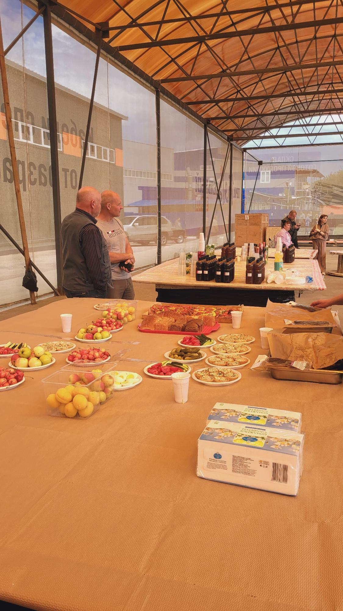 Two bald men stand talking under a covered outdoor structure with a translucent orange roof supported by metal beams. A table in front of them is laden with food and drinks. Several plates of fruit, including apples and cherries, are visible, as are containers of apricots. Other plates hold small pizzas and sliced boiled eggs. Behind these, a long platter holds various types of bread and pizza. Bottles of dark liquid, likely beverages, and plastic cups are also on the table. In the background, more tables are set up, and several people are visible, some interacting with food and drinks. A building with large lettering is visible behind the structure. The overall scene appears to be an outdoor gathering or buffet.