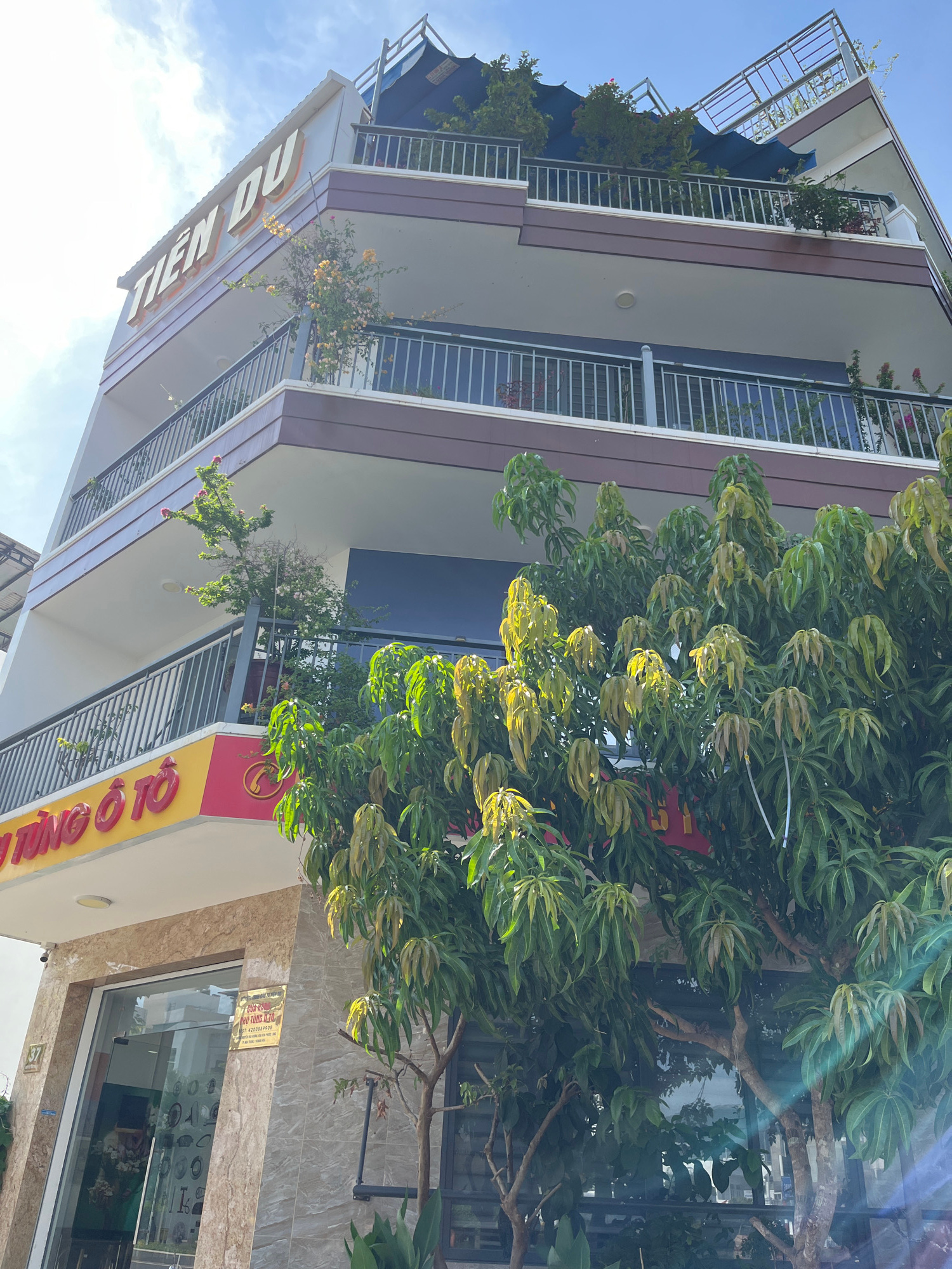 This low-angle, full-shot image captures a multi-story building under a bright, partly cloudy sky. The building, located in Nam Nha Trang Ward, Vietnam, has a modern design with multiple balconies. The upper facade displays prominent, raised, orange lettering that reads "TIÊN DU". Green railings line the balconies, some of which are adorned with potted plants and flowering vines. A mature tree with lush green leaves partially obscures the lower levels of the building. On the ground floor, a storefront with large glass windows is visible, featuring signage in Vietnamese. The overall scene is brightly lit, suggesting it is daytime, with some lens flare present in the lower right quadrant of the image.