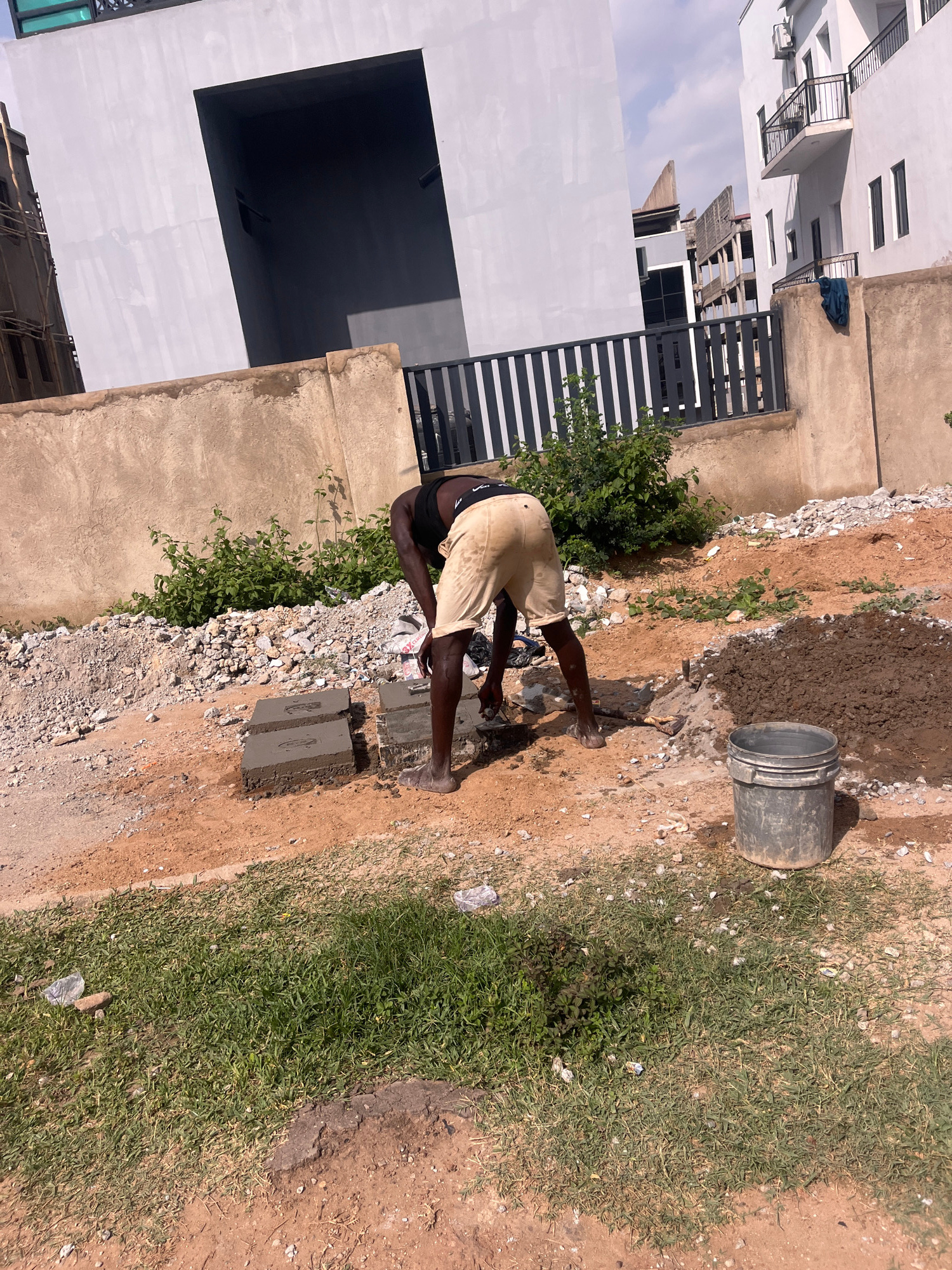 The image depicts a construction scene in Karum, Nigeria, under what appears to be a bright, sunny day. The main subject is a person, likely a construction worker, who is bent over, engaged in their work. They are wearing light-colored shorts and what appears to be dark underwear.
The setting is an outdoor construction site. In the foreground, a person is interacting with materials on the ground. There are several concrete blocks, some of which are wet, suggesting they are being prepared or have recently been used. A grey bucket, containing what looks like wet cement or mud, is positioned nearby. A small pile of gravel and debris is also visible. The ground is a mixture of dirt and patches of grass, indicating an unpaved area.
In the background, partially obscured by the construction activity, are elements of a more developed urban environment. A modern building with white walls, multiple windows, and balconies is prominent. A dark, open doorway or window is also visible on this building. Further back, other structures, some appearing unfinished with exposed scaffolding, are present, suggesting ongoing development. A black metal fence runs along a section of the site.
The activity taking place is clearly construction work, with the individual appearing focused on handling building materials. The time of day is likely midday, given the strong sunlight and shadows. The overall atmosphere is one of active labor in a developing urban area. No visible text is present in the image.