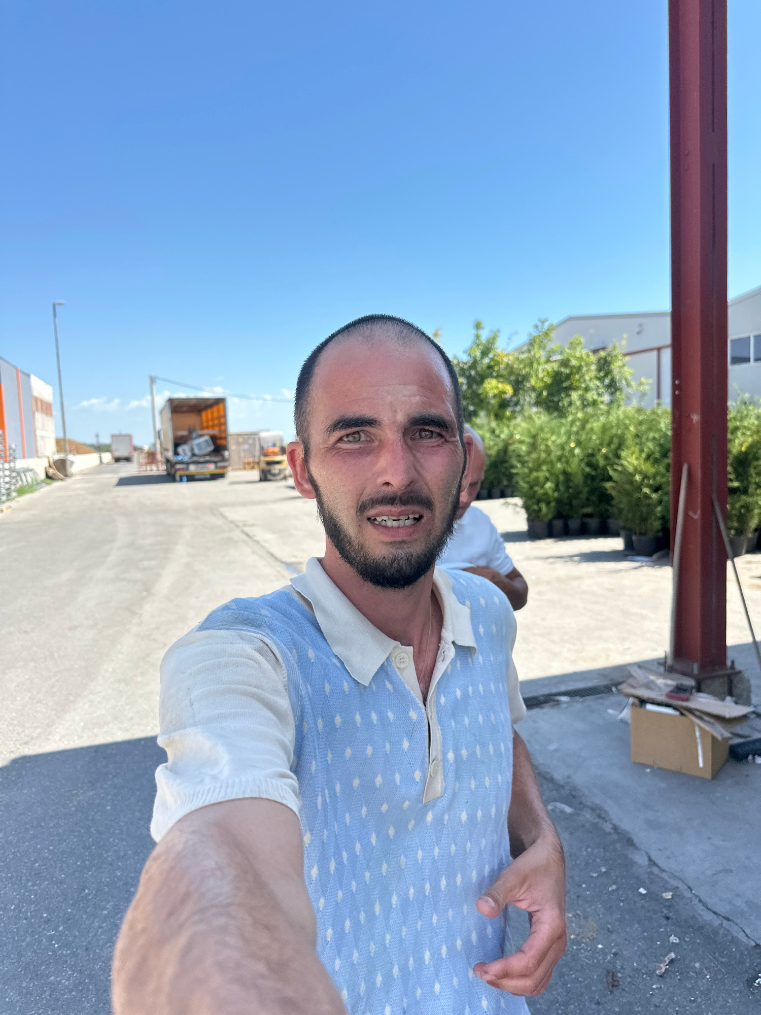 A close-up selfie of a man with short dark hair and a beard, wearing a light blue vest over a white collared shirt, is taken outdoors. He has braces on his teeth and his expression is neutral. In the background, a paved road leads towards industrial buildings under a clear blue sky. A truck with its back open is visible on the road, along with a forklift. Several potted green plants are arranged on the right side of the image, near a red metal pole. Another man in a white shirt is partially visible behind the main subject. The scene appears to be in an industrial or commercial area, potentially a loading dock or storage facility, in Rakića Kuće, Montenegro.