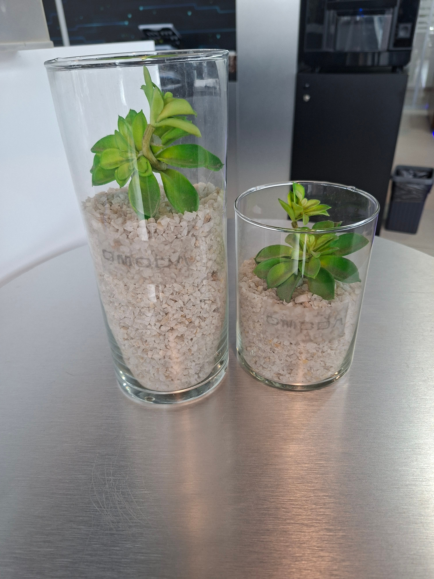 This image shows two glass cylindrical vases, each containing a small, artificial green succulent plant nestled in light-colored gravel. The vases are lying on their sides on a brushed metal surface, possibly a table or countertop. In the background, a dark, rectangular electronic device is visible, along with hints of what appears to be circuitry or a motherboard. The lighting suggests an indoor setting, likely during the daytime given the even illumination. There is some text imprinted on the glass of the upper vase, which reads "OMGD". The overall scene is minimalist and focuses on the decorative plants and their containers. The location context of Izhevsk, Russia, does not provide any specific visual cues within the image itself.