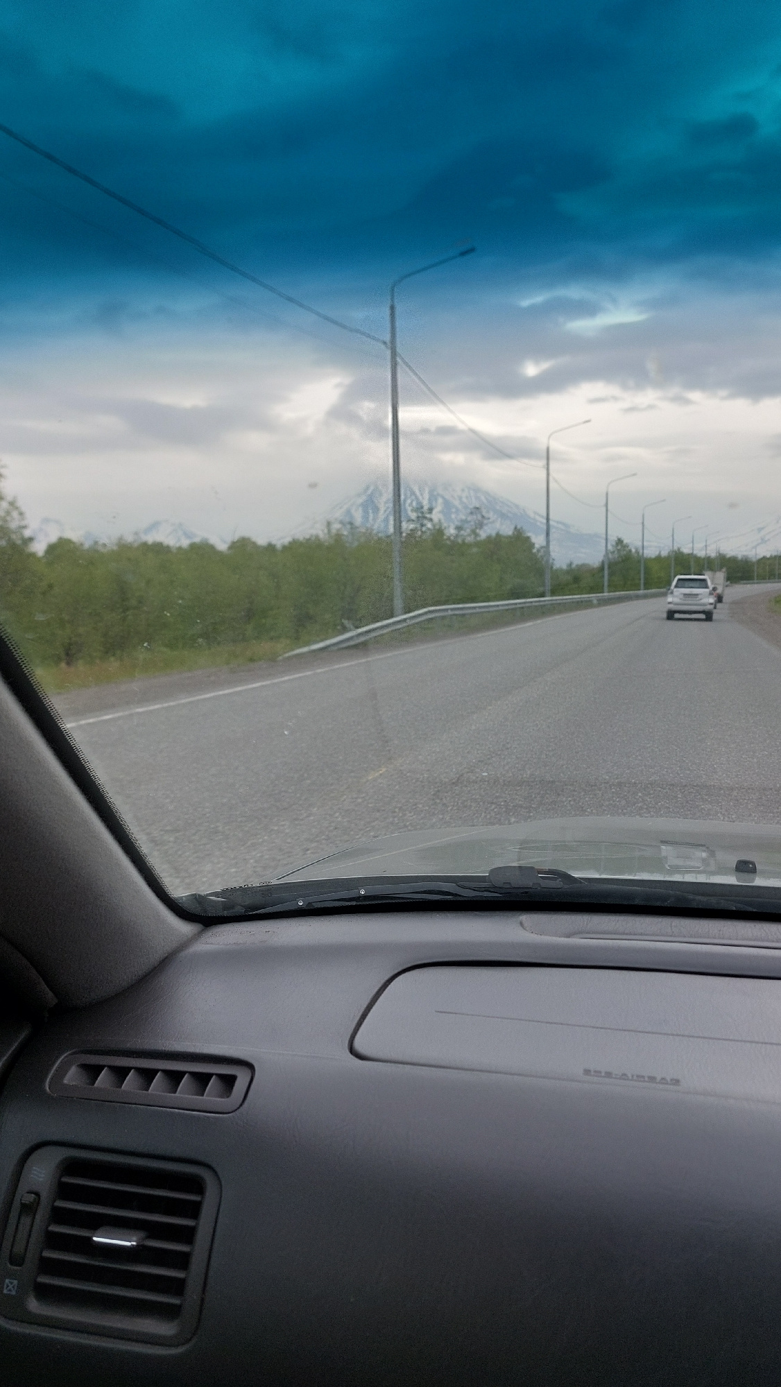 The image captures a view from inside a car looking out through the windshield and side window towards a road and a distant snow-capped mountain range under a cloudy, bluish sky. The car's dashboard and air vents are visible in the foreground. On the road, a white SUV is driving away from the camera, followed by another vehicle. Streetlights line the right side of the road, which is bordered by a guardrail and green trees. The scenery suggests a location in or near Yelizovo, Russia, a city known for its proximity to volcanoes.