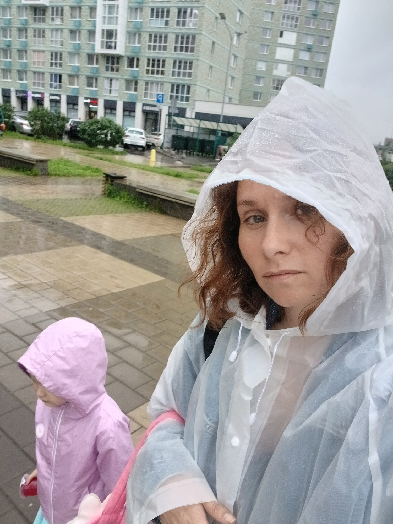 The image shows two people, likely an adult and a child, in what appears to be a city setting during a rainy day. The adult, wearing a white hooded poncho with water droplets on it, is looking towards the camera. Their face is partially visible, showing brown eyes and auburn hair. The child is wearing a pink hooded jacket and is positioned to the right and lower in the frame, facing away from the camera.
The background features a multi-story building with numerous windows, suggesting an urban environment. The ground is paved with interlocking bricks, with some grassy areas visible. The overall atmosphere is overcast, and the presence of rain gear indicates inclement weather. The scene captures a moment of everyday life, possibly a walk or an outing, being carried out despite the rain. The image is oriented sideways. No visible text is discernible.
