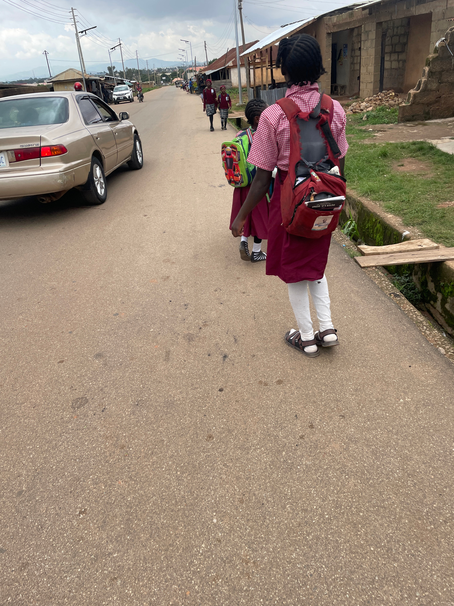 In Jos, Nigeria, a golden Toyota Camry is parked on the side of a paved road. Two young girls, presumably students, walk away from the camera on the same road. They are wearing matching maroon skirts and white socks, and one girl has a red and white checkered shirt. Both are carrying backpacks. The girl in the foreground has a red backpack with the words "PETER S. GONII" visible. The other girl carries a green backpack with a cartoon car design. Buildings, some appearing unfinished, line the right side of the road, along with a drainage ditch. A second white car and a motorcycle are visible further down the road, as well as other pedestrians. The sky is overcast.