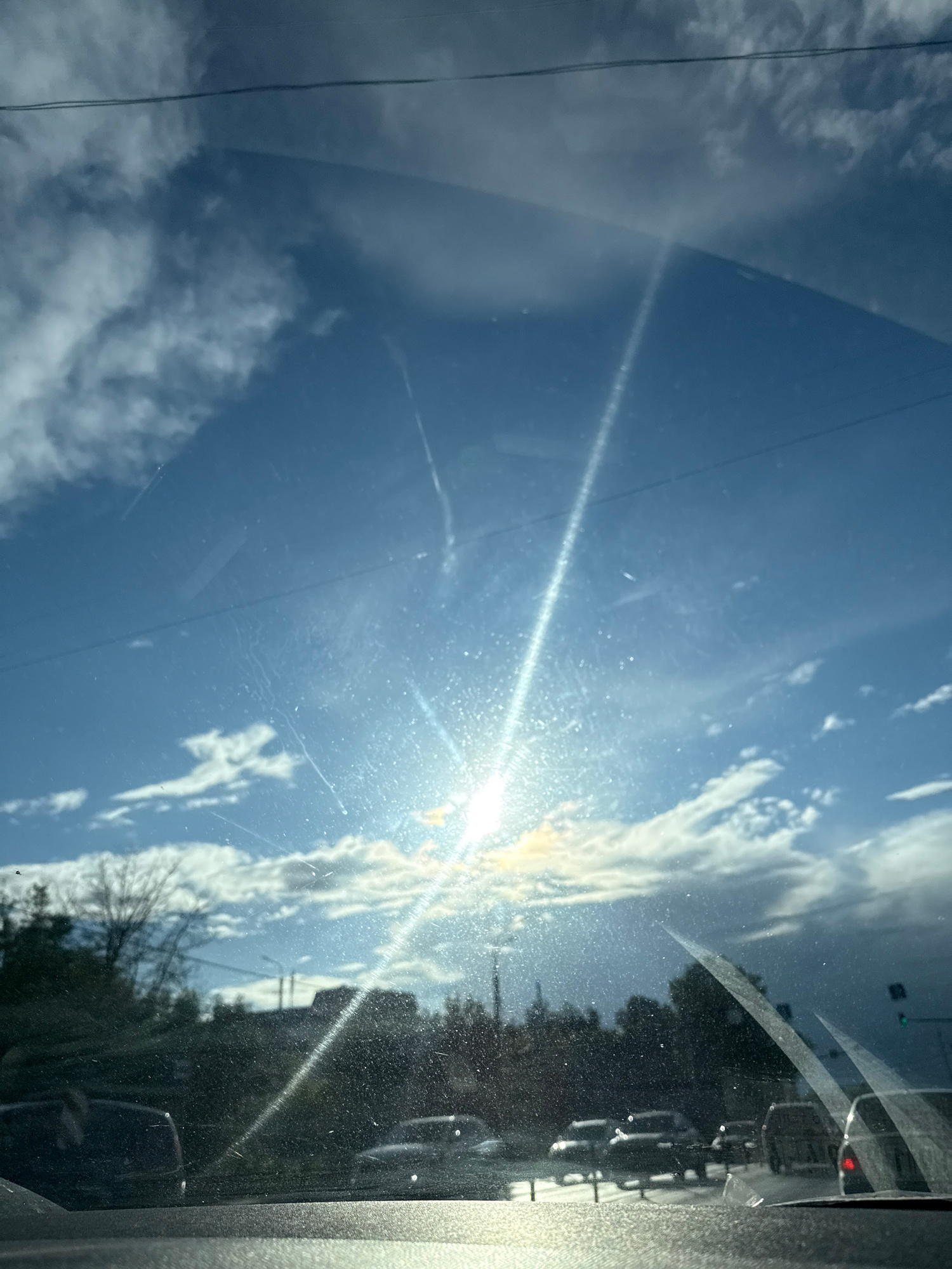 This media file depicts a reflection on a windshield, oriented vertically. The primary subject is a bright sun, positioned towards the right side of the frame and emitting a strong glare that creates a visible light streak across the glass. Above and to the left of the sun, white, fluffy clouds are scattered across a clear blue sky. The reflection also shows tree branches and some dark, silhouetted shapes that could be buildings or other vehicles. On the far right, a textured, gray surface suggests the interior of a vehicle, possibly the car's A-pillar. The presence of the sun and clouds indicates it is daytime, likely during fair weather. The reflection on the windshield, with the sun's glare and the captured sky, suggests the viewpoint is from inside a moving vehicle. The image is slightly dusty or smudged, with visible scratches on the glass. No people are directly visible, but the context implies someone is inside the vehicle. The scene appears to be outdoors, with natural light and elements of the sky.