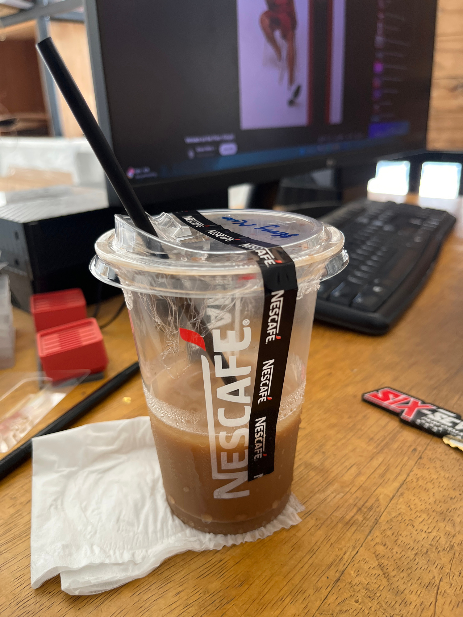 A close-up, eye-level, medium shot captures a disposable plastic cup filled with a brown beverage, likely iced coffee, resting on a white paper napkin on a wooden desk. A black straw extends from the cup at an angle. The cup is adorned with a black band featuring the "NESCAFÉ" logo repeated multiple times. In the background, a computer monitor displays an image, and a black keyboard is visible. To the left of the cup are two red, stacked objects, possibly small boxes. A black pen lies horizontally in the foreground to the left of the cup. A red and black sticker or keychain with the word "SIX" is positioned to the right of the cup, closer to the foreground. The lighting suggests an indoor environment, possibly an office or workspace. The scene implies a casual setting where someone is taking a break. The image does not provide specific clues to pinpoint the location in Kedawung, Indonesia.