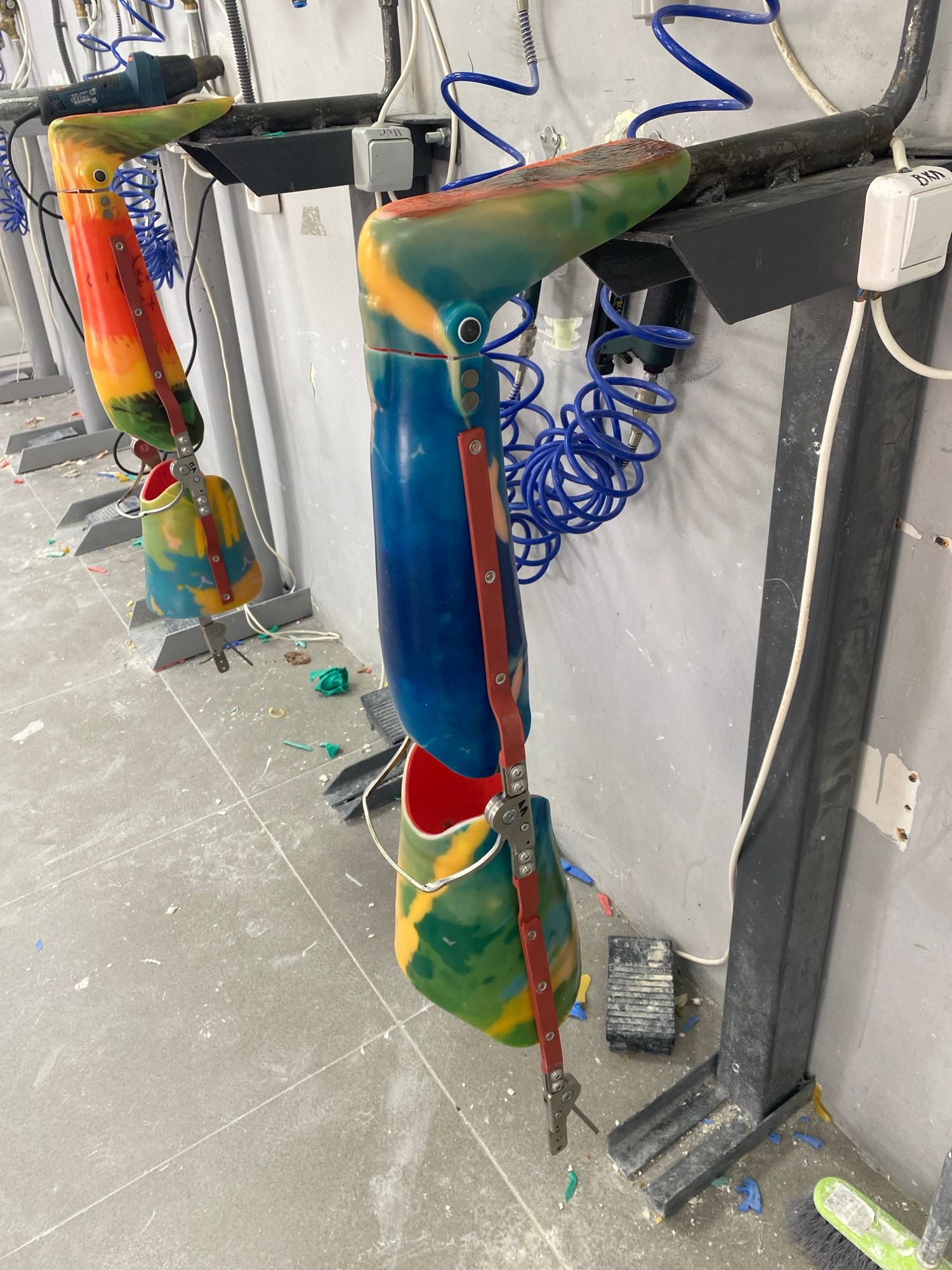 The image depicts a cluttered workshop or laboratory setting. In the foreground and midground, several colorful prosthetic limbs, possibly for legs, are hanging from black metal racks mounted on a light gray wall. These prosthetics are made of a marbled material in shades of orange, yellow, green, blue, and red, and appear to be in various stages of assembly or repair, with exposed mechanical components and red straps. Blue coiled air hoses are visible, attached to some of the equipment. The floor is made of gray tiles and is littered with debris, including small plastic pieces, scraps of material, and dust, suggesting an active work environment. To the right, a dark gray vertical metal support structure is visible, with a white electrical cable running along it. In the background, other similar prosthetic limbs and equipment are partially visible. The overall impression is one of a functional, albeit messy, workspace for the creation or maintenance of prosthetic devices. The image does not provide any specific contextual information that would allow for its location to be identified as Vladimir, Russia, or any other specific city.