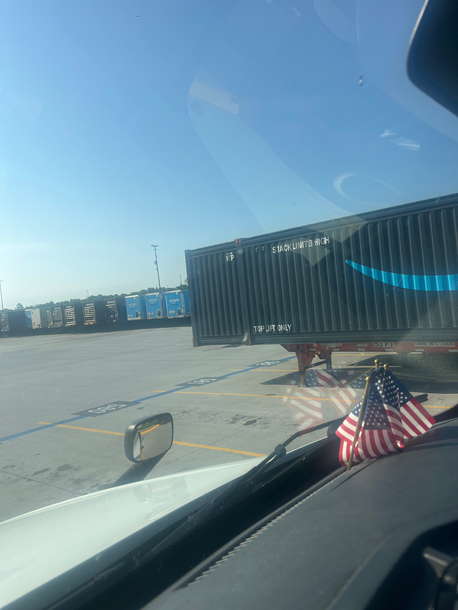 The image, taken from inside a vehicle, shows a clear blue sky with a few wispy clouds and a distant object that may be an airplane. The foreground is dominated by the dashboard and windshield of the vehicle, with two small American flags attached to the dashboard. A side mirror is visible on the left. The background reveals a vast outdoor lot filled with parked semi-trailer trucks. Many of these trailers bear the Amazon logo. One large black shipping container with the Amazon logo prominently displayed is in the mid-ground. Text on the container reads "STACK LIMIT 8 HIGH" and "TOP LIFT ONLY". Parking lot markings, including yellow and blue lines and numbered spots, are visible on the ground. The overall scene appears to be a logistics or distribution center in Pooler, United States of America.