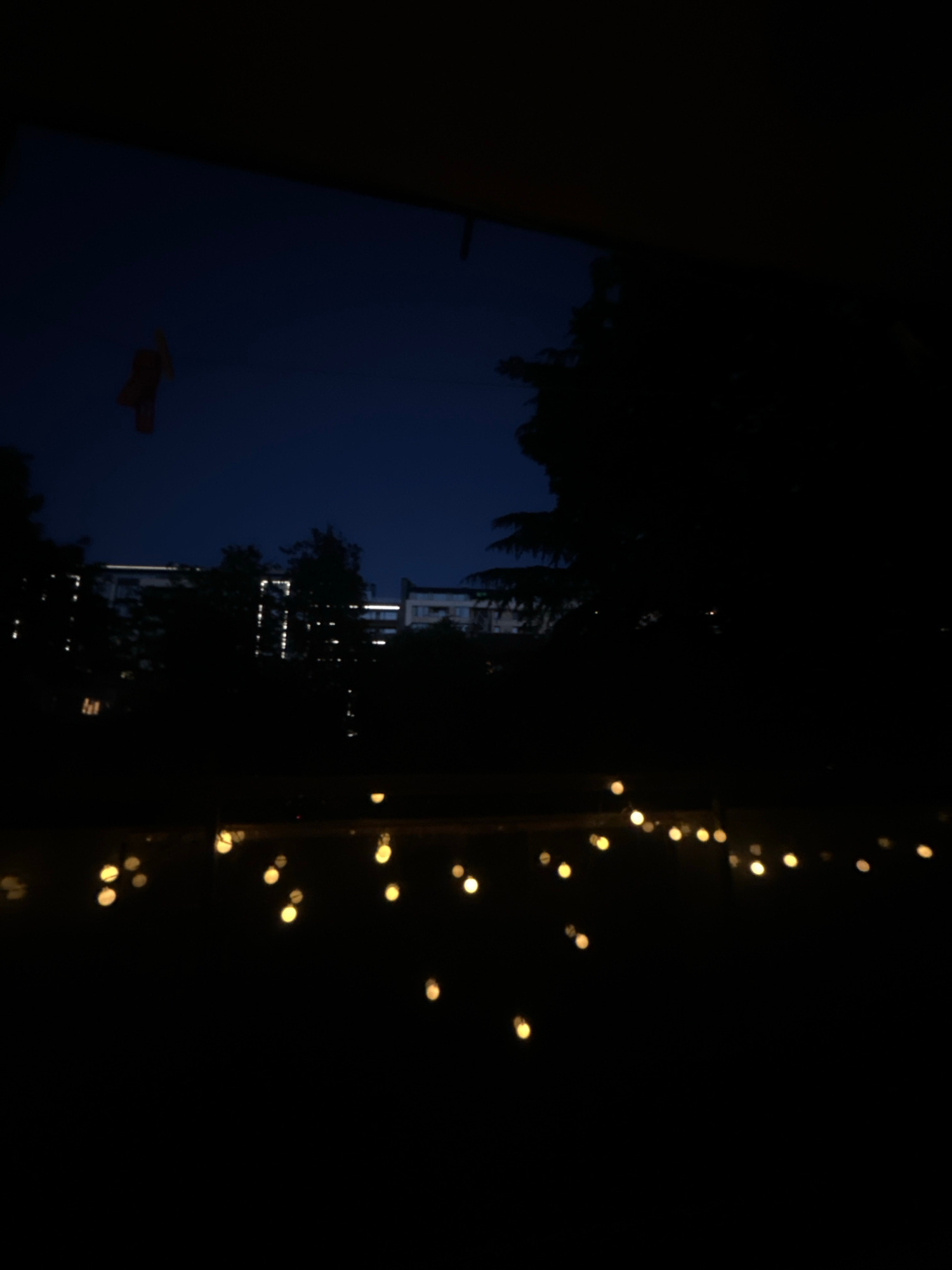A nighttime view from a window shows a dark blue sky above the silhouettes of trees and buildings. In the upper left corner, a small, reddish-orange object, possibly a kite or decoration, hangs against the sky. In the middle ground, the illuminated windows of apartment buildings create bright, linear patterns against the dark structures. In the foreground, a string of yellow lights, blurred and out of focus, hangs low, casting a warm glow against the darkness. The scene appears to be an urban setting, potentially a residential area in Sofia, Bulgaria, as suggested by the architecture and the presence of trees and lights.