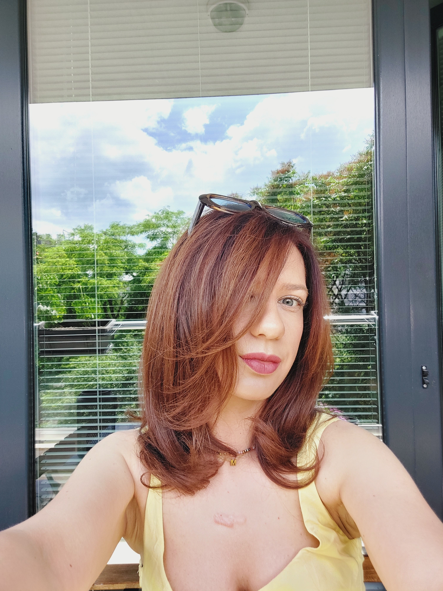 A medium shot captured from a slightly low angle features a person with shoulder-length reddish-brown hair, styled with layers and bangs partially obscuring the right side of their face. The person is looking towards the camera, with their left eye visible. They are wearing a light yellow top and a necklace with a pendant. Sunglasses are resting on top of their head. The background consists of a large window with horizontal blinds, revealing lush green trees and a cloudy blue sky. The setting appears to be outdoors, possibly a balcony or patio, overlooking a residential area in Sofia, Bulgaria, with dense foliage characteristic of the city's many green spaces. The lighting suggests a daytime scene, likely in the late morning or early afternoon, with natural light illuminating the subject and the surroundings.