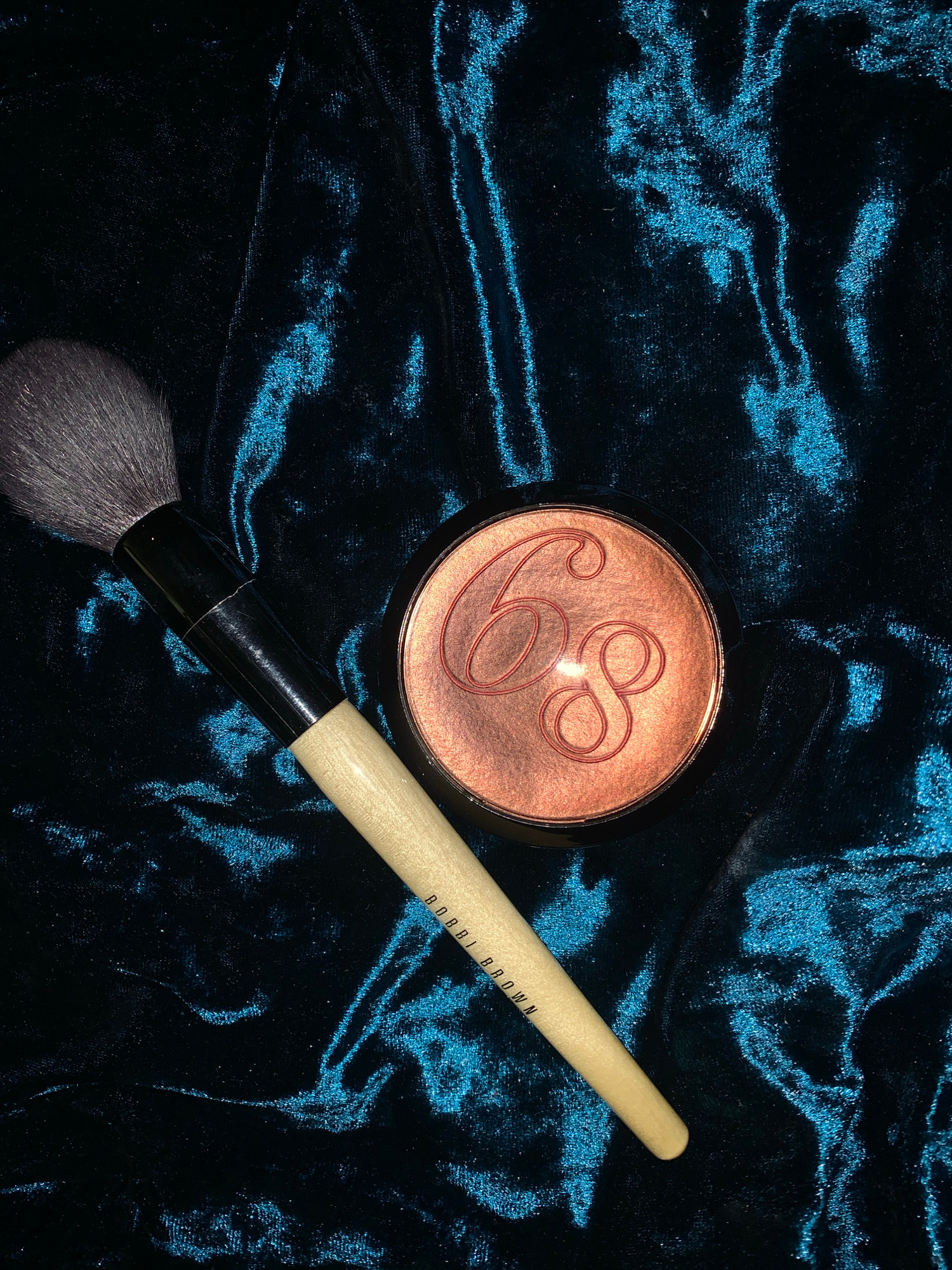A makeup brush and a compact of bronzer are laid out on a dark, teal-colored velvet fabric. The brush has a light-colored handle with "BOBBI BROWN" printed in black text. The bronzer compact is round with a copper-toned powder inside, embossed with a stylized "68". The velvet fabric has a high sheen and appears to be draped, creating folds and highlights.