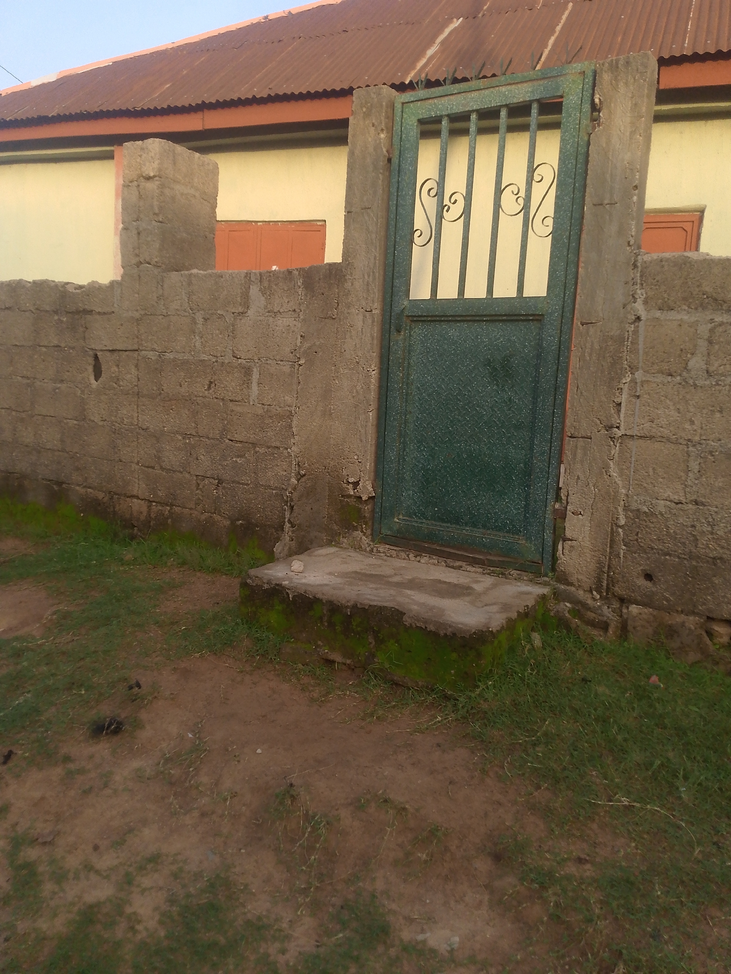 The image shows a green metal gate with decorative scrollwork in its upper half, set into a concrete and cinder block wall. The gate is partially open and leads to a building with a yellow facade and a corrugated metal roof. The wall is made of rough, textured concrete blocks, with a small concrete step leading up to the gate. To the left of the gate, a section of the wall is incomplete, revealing more of the yellow building wall behind it. The ground in front of the wall is a mix of dirt and patchy green grass. The sky is visible in the upper portion of the image, appearing light blue and clear.