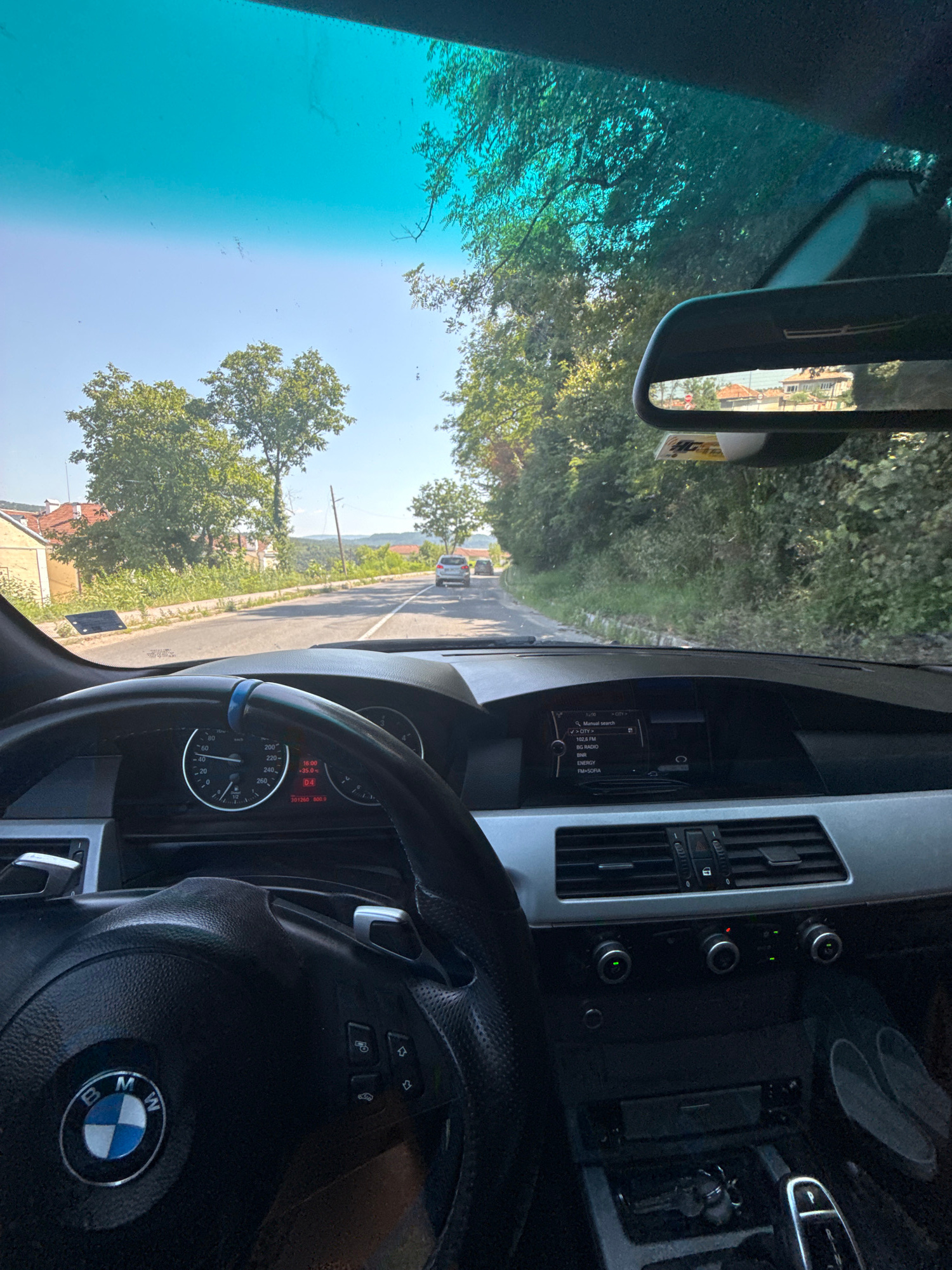 The image shows the interior of a BMW car, likely on a road in or near Dryanovo, Bulgaria. The view is from the driver's perspective, looking forward through the windshield and at the dashboard. The dashboard features a steering wheel with the BMW M logo, a speedometer, and a central infotainment screen displaying radio information and navigation prompts. Outside the car, a paved road is visible, bordered by green trees and vegetation. Houses with red-tiled roofs can be seen in the distance on the left side of the road. Another car is ahead on the road. The sky is clear and blue. The scene suggests a daytime drive through a semi-rural area.