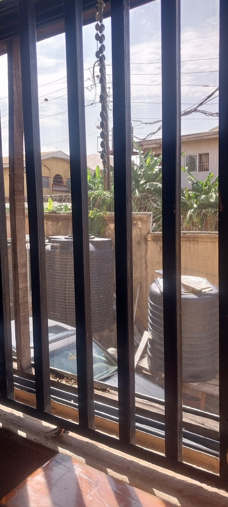 The image shows a view through a window with horizontal bars and a screen. The scene outside appears to be a residential area. In the background, there are buildings with light-colored walls and some greenery, possibly trees or large plants. Overhead, there are electrical wires strung across the sky, suggesting an urban or semi-urban environment. The lighting indicates it is daytime, with bright sunlight casting shadows. The overall impression is a glimpse into a typical street view in Onitsha, Nigeria. The presence of the window bars and screen implies a sense of security or protection.