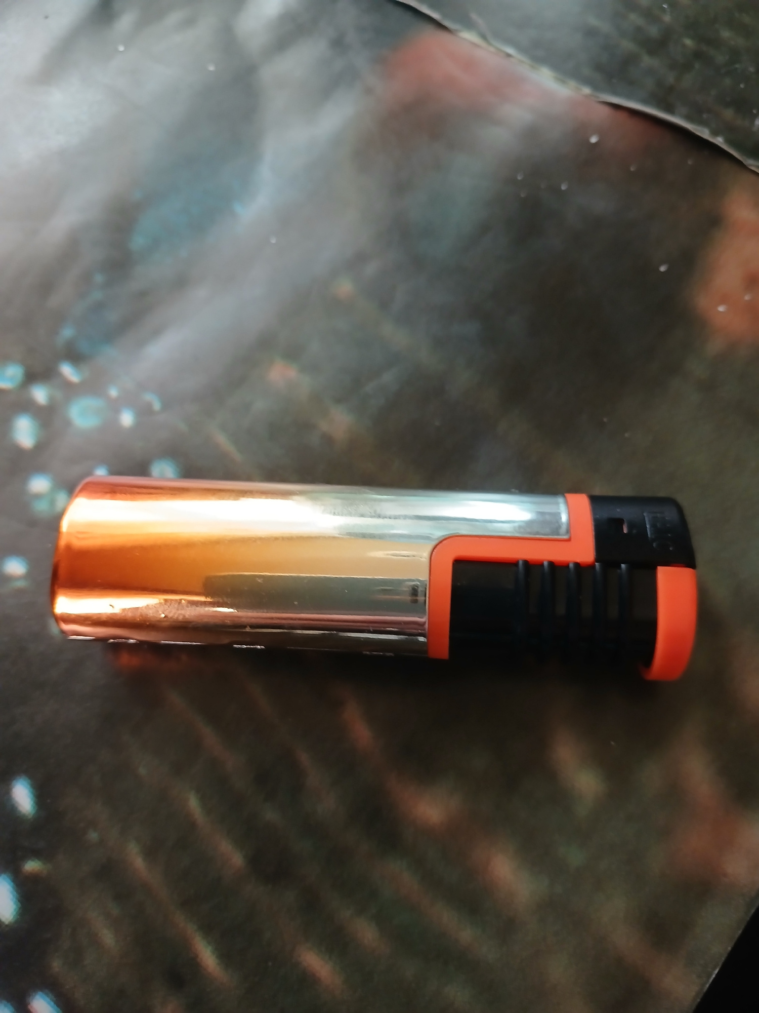 The image shows a single object, a butane lighter, lying horizontally on a patterned surface. The lighter has a metallic orange-toned body and a black and orange plastic base. The background surface is dark with blurred iridescent and reddish-brown splotches, possibly a printed cloth or mat. There are no people or other objects visible in the scene. The lighting appears to be from above, casting a subtle shadow. The location of this scene in Hlushky, Ukraine, is not discernible from the provided image.