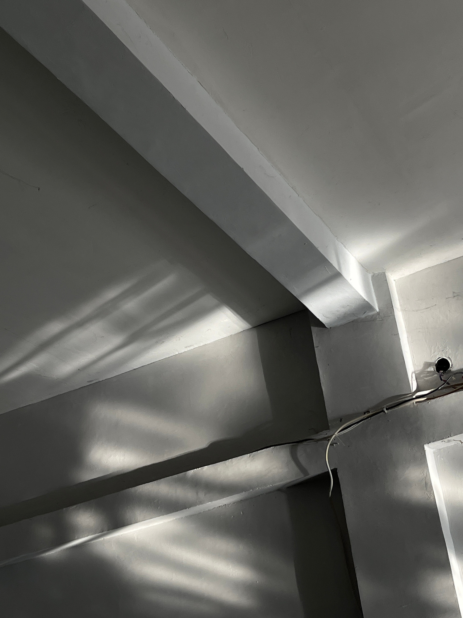 The image shows a close-up, low-angle interior view of a building under construction or renovation in Kedawung, Indonesia. The scene is dominated by concrete beams and walls, rendered in shades of gray and white. Sunlight streams in from an unseen source, casting long, parallel shadows that create a dramatic, striped pattern across the surfaces. These shadows suggest that the light is originating from a barred window or external structure.
Several structural elements are visible, including a prominent horizontal beam running diagonally across the upper portion of the frame, a smaller beam or ledge below it, and vertical corners where walls meet. The surfaces appear unfinished, with visible texture and some imperfections. Electrical conduits and wires are present along one of the walls, with a circular opening suggesting an electrical junction box or outlet. The overall impression is one of a utilitarian, possibly industrial or unfinished space, illuminated by natural light.