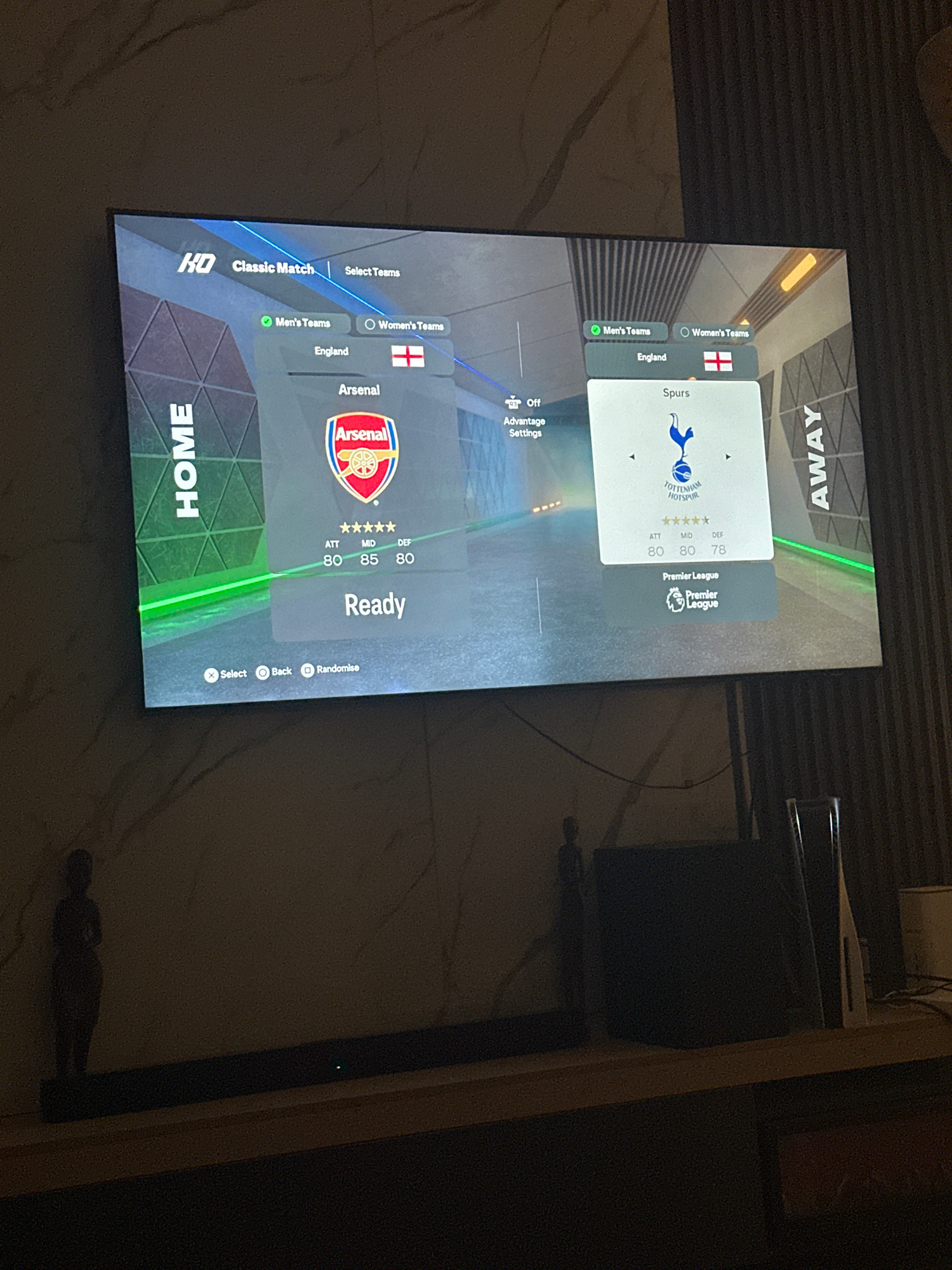 The image shows a television screen displaying a video game interface, likely a soccer game, with two teams selected. The left side of the screen shows "HOME" with the Arsenal logo and team statistics. The right side of the screen shows "AWAY" with the Tottenham Hotspur (Spurs) logo and team statistics. The game appears to be set in England, as indicated by the flags and league displayed. Below the team selections, there are options to "Select", "Back", and "Randomise". The television is mounted on a wall with a patterned finish. The room appears to be a living area, with a wooden shelf below the television on which several objects are placed, including what appears to be a soundbar and two small decorative figurines. The lighting in the room is dim, with some light emanating from the television screen and a few visible light fixtures. The overall scene is consistent with a home entertainment setup. The mention of Gwarinpa, Nigeria, is a contextual detail that cannot be visually confirmed from the provided image alone, but it suggests the location where this scene is situated.