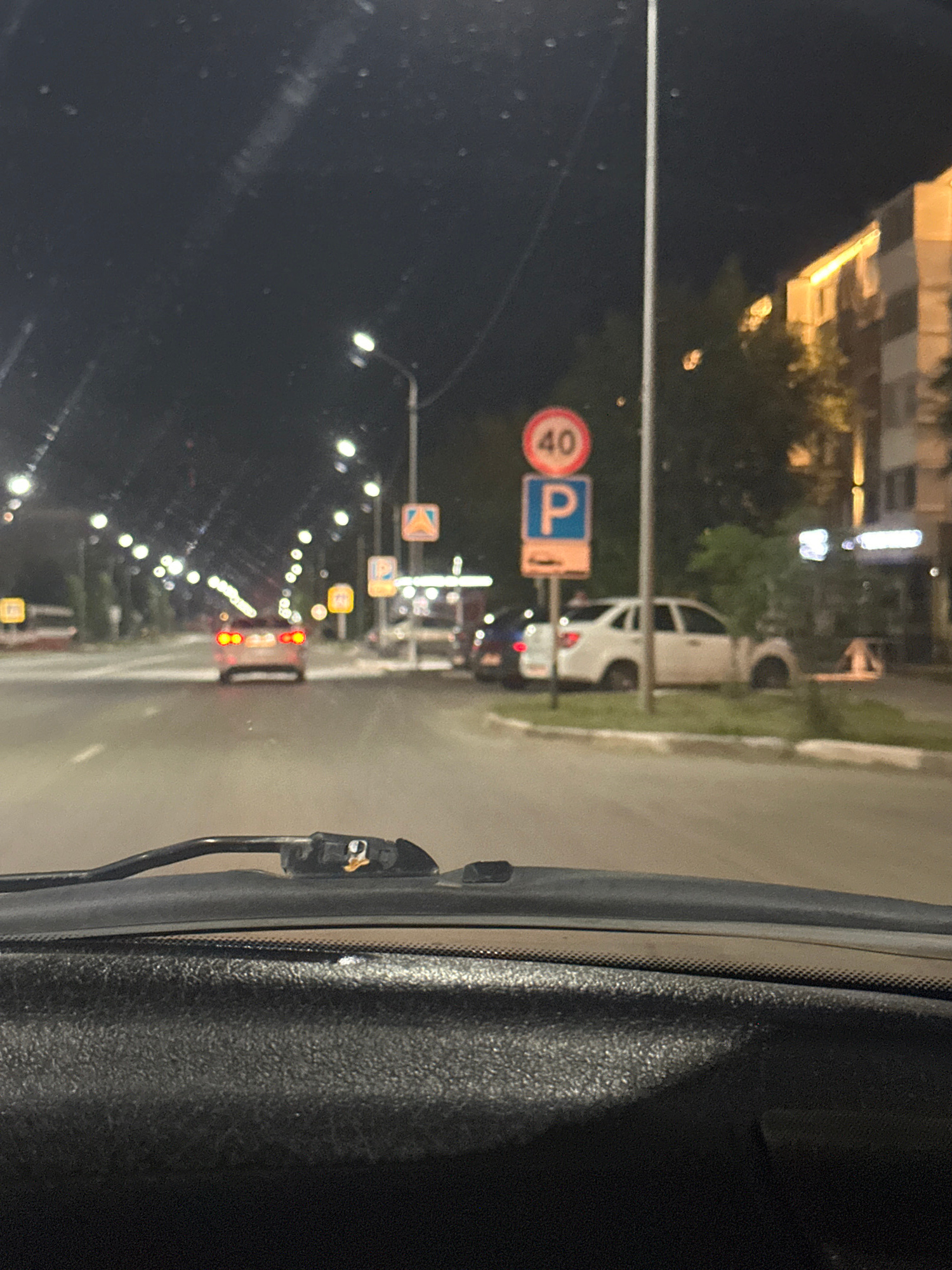 The image shows a night-time view from inside a car, looking out through the windshield. The scene depicts a street with lampposts illuminating the road and sidewalk. There are several traffic signs visible, including a speed limit sign indicating "40" and a parking sign with a blue "P". Other signs with orange and white geometric patterns are also present.
Several vehicles are on the road, including a white SUV in the background and what appears to be a truck or bus with red taillights further down. The overall impression is of a moderately busy street in an urban or suburban area at night. The lighting suggests it is nighttime, and the lack of visible precipitation indicates fair weather. The presence of buildings in the background, though blurred, hints at an urban setting. The photo's orientation is rotated 90 degrees clockwise. There is no discernible human activity beyond the implied presence of the driver and passengers within the vehicle from which the photo is taken. The text on the signs is limited to numbers and symbols.