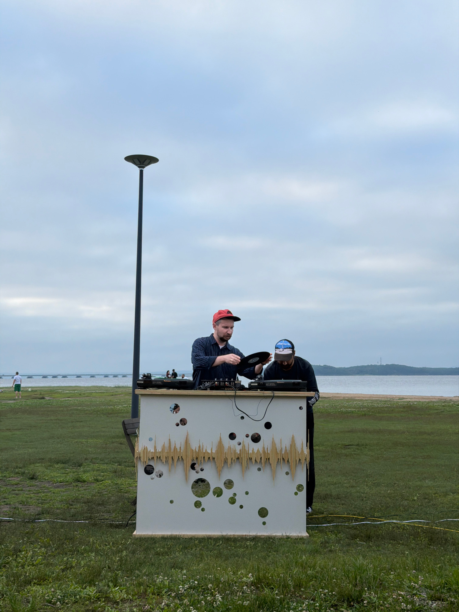 Two DJs are operating a sound system on a table with a decorative front panel in an outdoor setting. The DJ on the left, wearing a red baseball cap and a striped shirt, is holding a vinyl record. The DJ on the right, wearing a blue and black baseball cap and a dark shirt, is focused on the equipment. They are positioned in front of a body of water, likely the Sea of Japan, with a bridge visible in the background. The environment is grassy with a few scattered trees and a lamppost. The sky is overcast. Based on the context of outdoor DJ setups near the sea and bridges, this scene could be in Vladivostok, Russia, specifically along the coast or near one of its many waterfront parks.