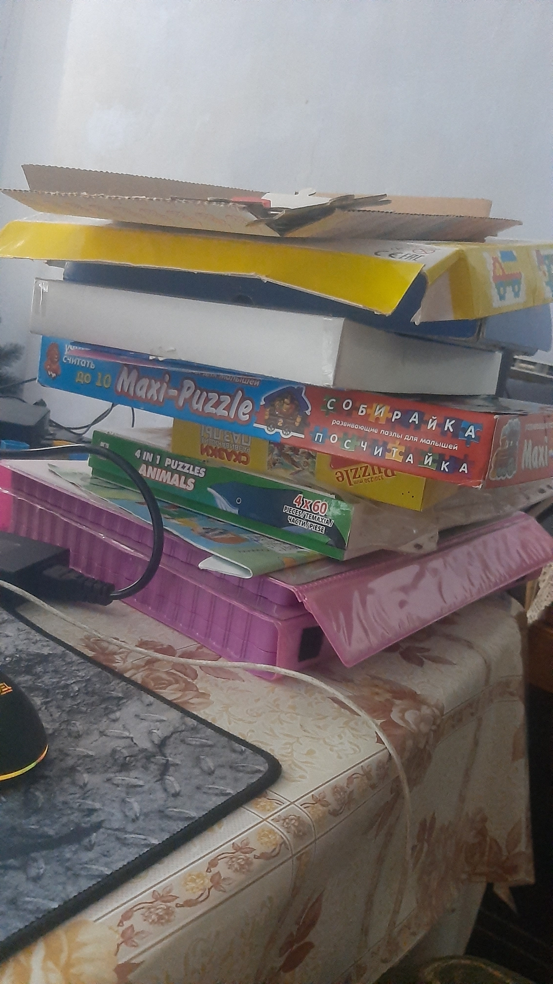 In a cluttered indoor environment, a stack of puzzle boxes sits on a table covered with a floral-patterned tablecloth. The table is located in the city of Katlabuh, Ukraine. The stack includes a blue box labeled "Maxi-Puzzle" with Russian text, a green box for "4 IN 1 PUZZLES ANIMALS," and other boxes with visible Russian text including "СОБИРАЙКА" and "ПОСЧИТАЙКА". A black computer mouse pad with a metallic texture is positioned to the left of the table. The background is a plain, light-colored wall. The lighting suggests an indoor setting with artificial light. There are no people visible in the image.