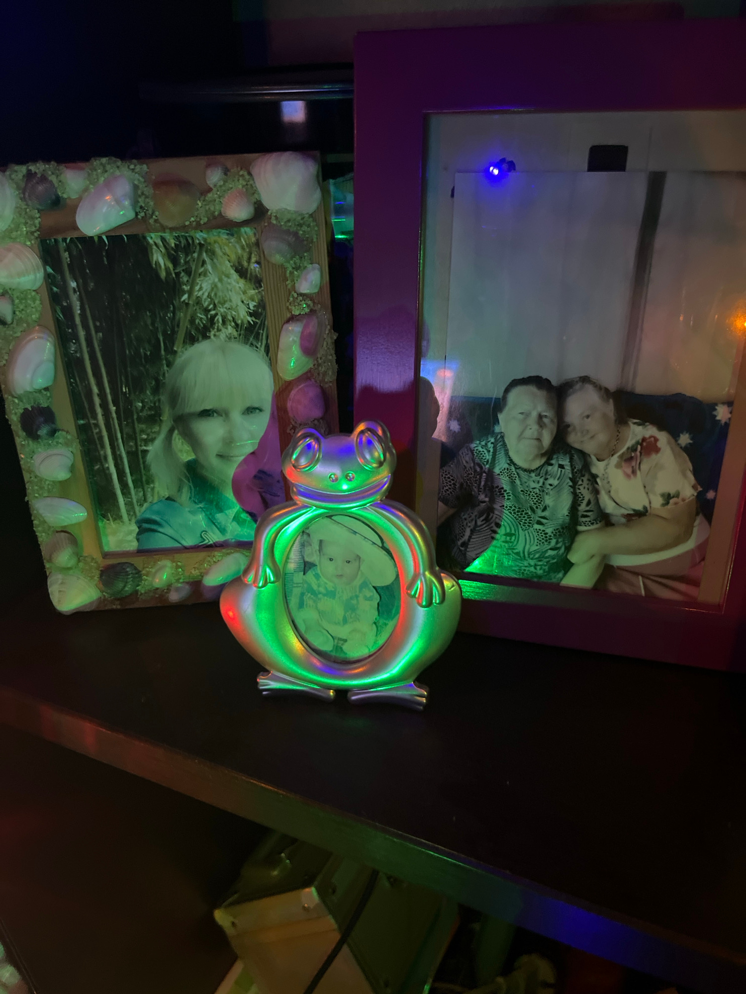 The image shows a shelf with three framed pictures and a small frog-shaped frame with a picture inside. On the left, a framed picture with a seashell border shows a blonde woman with her hair in a ponytail, smiling. The frame is decorated with various seashells and what appear to be small stones. To the right of this is a larger, purple-framed picture that contains a photo of two elderly women. The woman on the left is wearing a patterned shirt, and the woman on the right is wearing a floral print shirt and has her arm around the other woman. A small, frog-shaped frame sits in front of the two larger frames. This frame has a metallic, iridescent finish and is illuminated by colorful lights. Inside the frog frame is a picture of a baby. The overall scene is dimly lit with colored lights reflecting off the objects. There is no indication of the city of Kazan, Russia in the image itself.