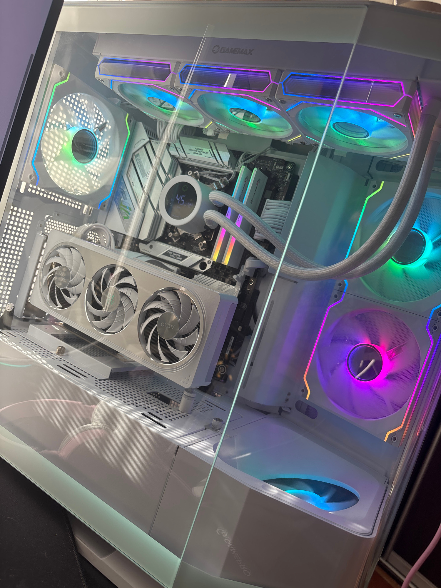 A white computer case with multiple RGB fans is shown. The computer case has a glass side panel, revealing the internal components. A graphics card with three fans is installed horizontally. The computer has RGB lighting on the fans and other components, displaying various colors including green, blue, purple, and pink. There is a circular display on the motherboard showing "45". The background is dark, and the scene is illuminated by the RGB lighting of the computer. The image appears to be taken indoors, likely in a home or office setting.