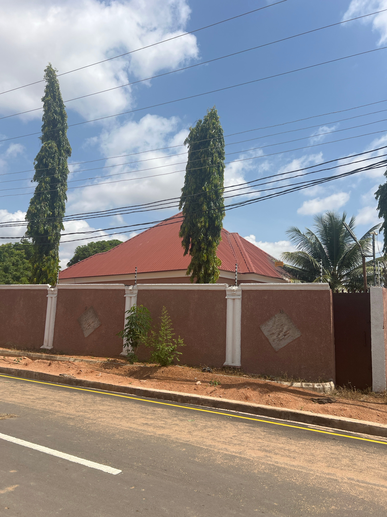 The image depicts a residential scene in Howayi, Nigeria, under a clear, blue sky with scattered white clouds, suggesting it is daytime. The primary subjects are a house with a red, corrugated metal roof and a brown, stuccoed wall topped with decorative white pillars and diamond-shaped accents. A tall, slender tree with dense green foliage stands prominently in the foreground, next to another similar tree. Electrical wires crisscross the sky above, casting shadows. The house is bordered by a sandy embankment and a paved road with a yellow line and a white lane marking. A palm tree is visible in the background, hinting at a tropical or subtropical climate. The scene is peaceful and static, with no people or discernible activity, conveying a sense of quiet residential life. There is no visible text in the image.