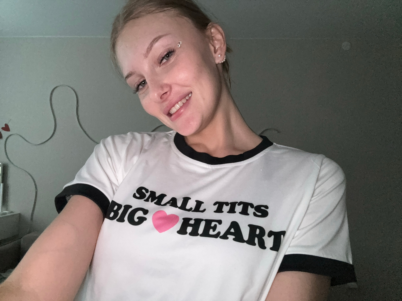 A young woman with blonde hair pulled back is smiling at the camera. She is wearing a white t-shirt with black trim around the collar and sleeves. The t-shirt has black text on it that reads "SMALL TITS BIG HEART" with a pink heart in place of the word "BIG". She has several small silver piercings on her face, including one above her eyebrow and others on her cheekbone. She also has earrings. The background is a lightly colored wall with some faint, dark grey wavy lines and a few small red heart shapes painted on it. The lighting is dim. The image appears to be a selfie taken indoors, possibly in a bedroom. There is no indication of the location being the city of Perm, Russia.