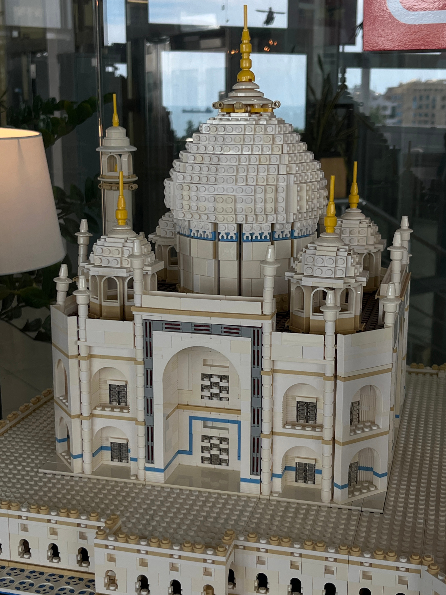 A detailed LEGO model of the Taj Mahal is displayed on a grey LEGO baseplate. The model features a large central dome, surrounded by four smaller domes and four minarets. The exterior of the model is primarily white, with accents of blue and gold. The model is placed in what appears to be an indoor setting, with reflections of the outside world visible in the glass enclosure. A lampshade is visible to the left of the model, and greenery is present in the background. The specific location within Yalta, Ukraine, is not discernible from the image.