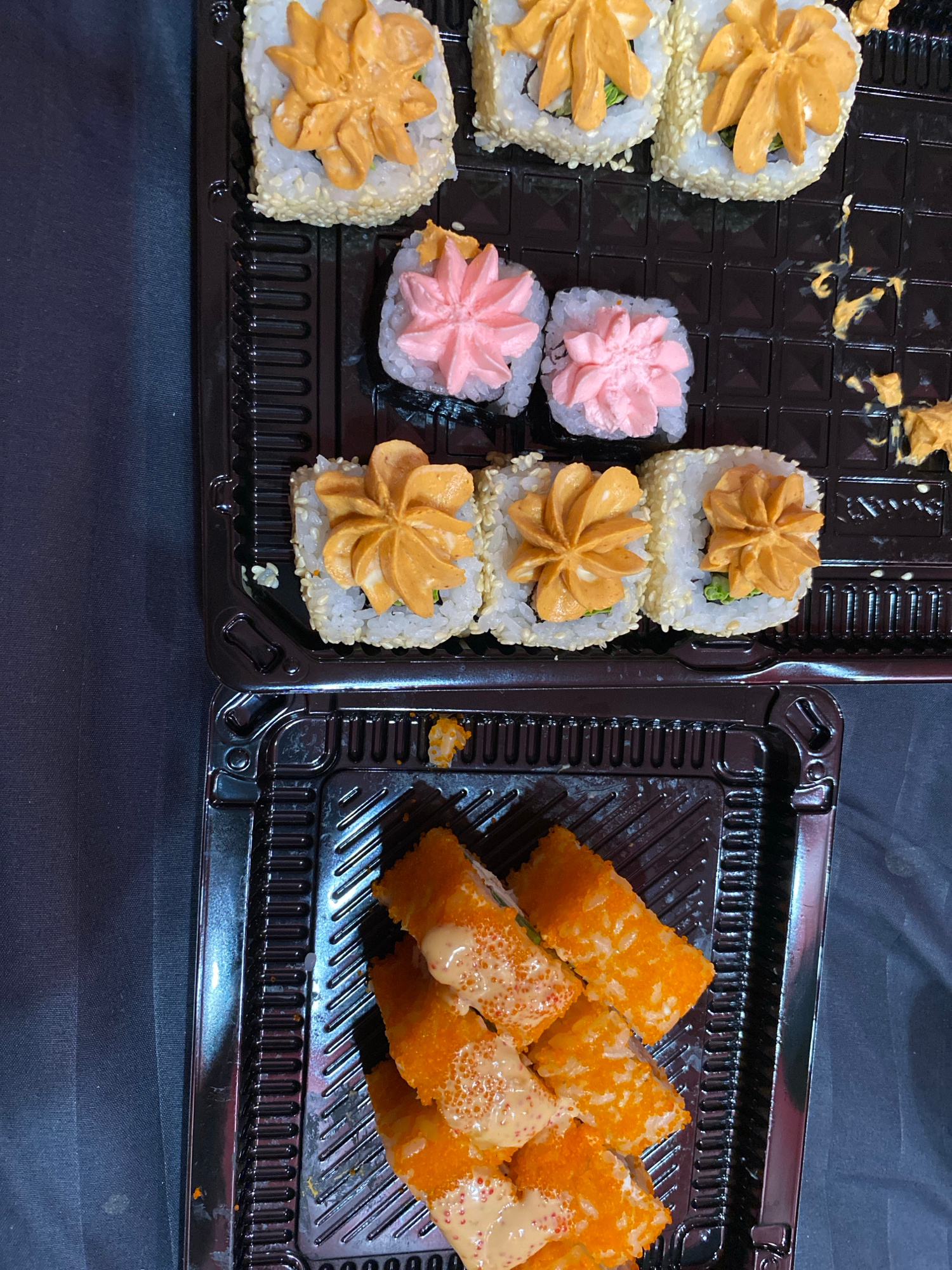 A top-down, medium shot captures a black plastic takeout container filled with sushi. The container is divided into two sections. The top section holds six sushi rolls arranged in two rows of three. The rolls are topped with a creamy, orange sauce piped in a star-like pattern, and the outer edge of the rice is coated with white sesame seeds. Below these, in the same section, are two sushi rolls topped with a pink cream piped in a similar star pattern. To the right of these rolls, there are remnants of orange sauce.
The bottom section of the container contains several pieces of sushi rolls that are cut into rectangular pieces. These pieces are coated in small, orange tobiko (flying fish roe) and drizzled with a creamy, beige sauce.
The container rests on a dark, textured surface that appears to be fabric, possibly a tablecloth or a dark cloth backdrop. The lighting is diffused, casting soft shadows. The image quality is slightly grainy.
The scene is located in Yerevan, Armenia.