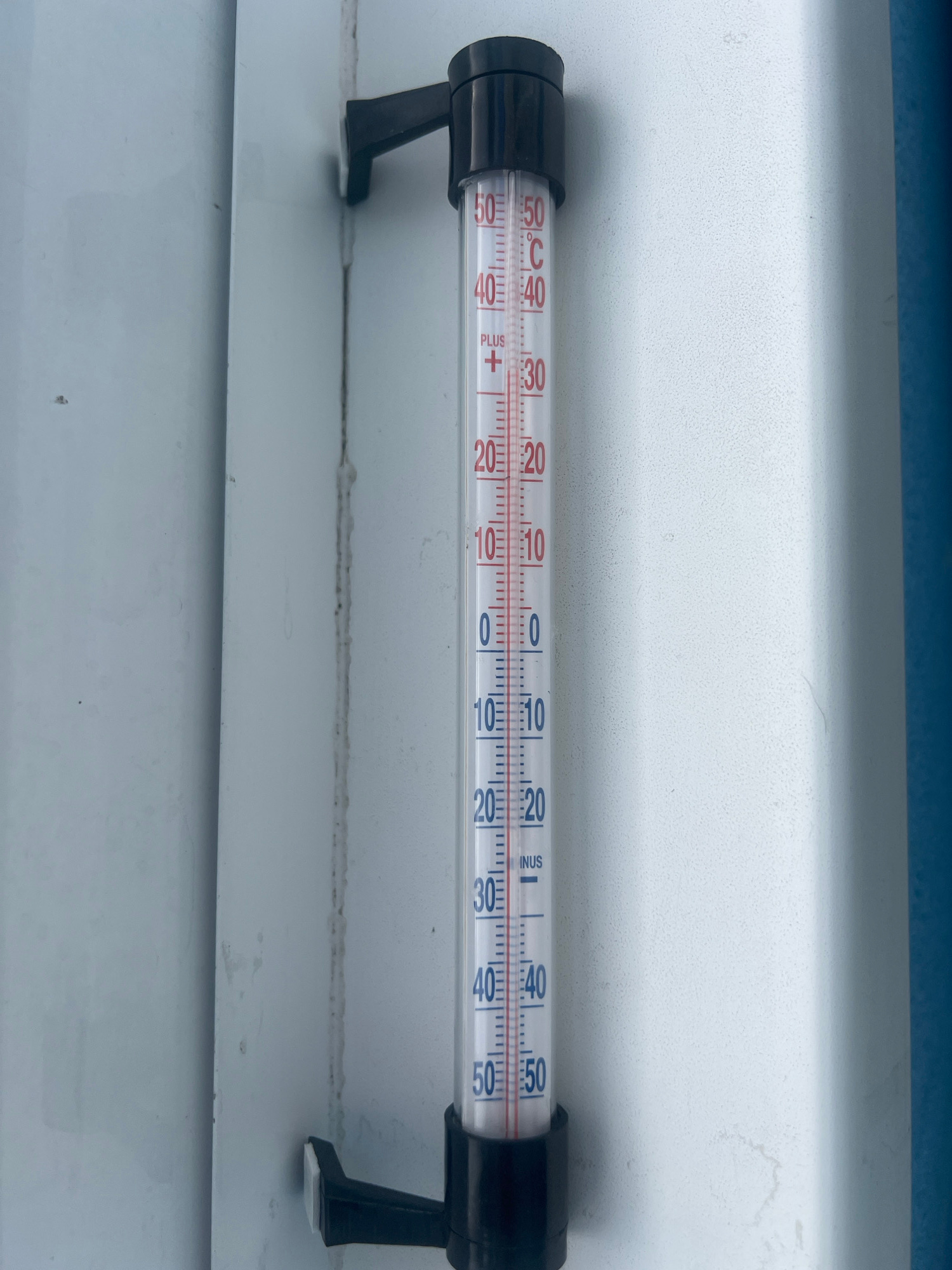 A horizontal thermometer is mounted on a textured white surface, likely an outdoor wall or window frame. The thermometer is enclosed in a clear glass tube with black plastic mounting brackets on either end. The markings on the thermometer range from -50 to 50 degrees Celsius, with red markings for positive temperatures and blue markings for negative temperatures. The bulb of the thermometer is on the left side, and the liquid column is visible. The liquid column is slightly above the 0-degree mark, indicating a temperature slightly above freezing. There is text visible on the thermometer, including "PLUS" and "MINUS", as well as the degree symbols "°C" and "°F". The lighting suggests it is daytime, and the overall scene appears to be outdoors. The background consists of a blue band at the top and the white textured surface where the thermometer is attached, with horizontal lines suggesting the surface is made of overlapping panels. The scene is static, showing an object in its setting.