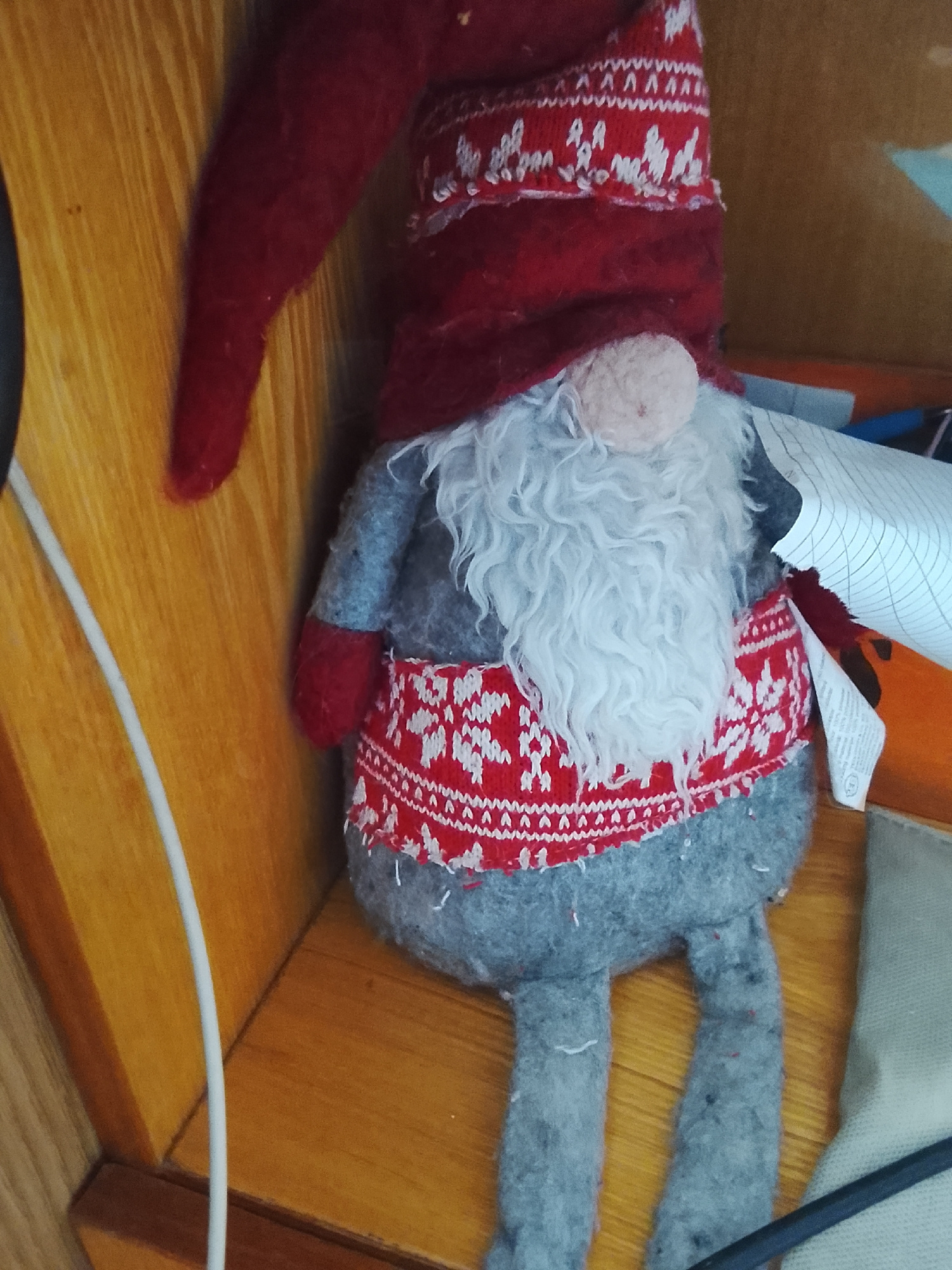 A stuffed gnome with a red hat, white beard, and a red and white patterned tunic stands on a wooden shelf. The gnome is positioned slightly off-center to the right, with its pointed red hat tilted to the left. Its white beard is fluffy and fills the lower portion of its face. The gnome wears a gray tunic and a red and white patterned belt or apron. Its legs, made of gray fabric, hang down. To the left of the gnome, a white cable runs vertically down the wooden surface. In the background, the wooden shelf continues, and to the right, some papers with grid lines are visible, along with a hint of orange. The lighting suggests an indoor environment. There are no people or animals present in the image. The image does not provide any information about its location in the city of Almaty, Kazakhstan.
