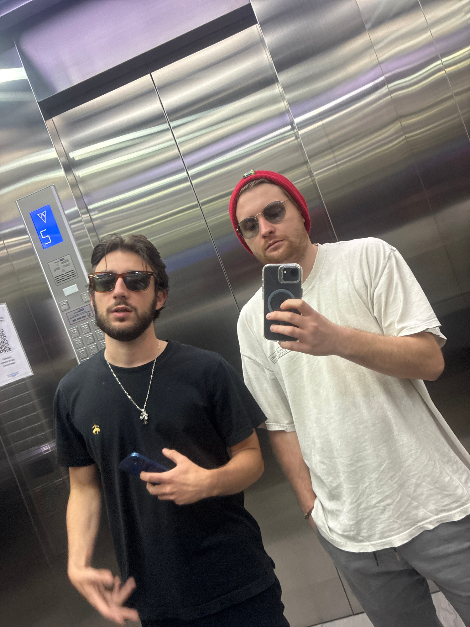 The image shows two young men in an elevator, taking a selfie. The elevator has brushed metal walls and a control panel with buttons and a digital display. The man in the foreground is wearing a black t-shirt, sunglasses, and a silver necklace with a cross pendant. He is holding a blue smartphone. The man behind him is wearing a white t-shirt, a red beanie, and sunglasses. He is holding a black smartphone. The lighting suggests it is daytime. The men appear relaxed and are posing for the photo. There is some text on the elevator control panel, but it is not clearly legible. The setting appears to be a modern building in Huixquilucan de Degollado, Mexico.