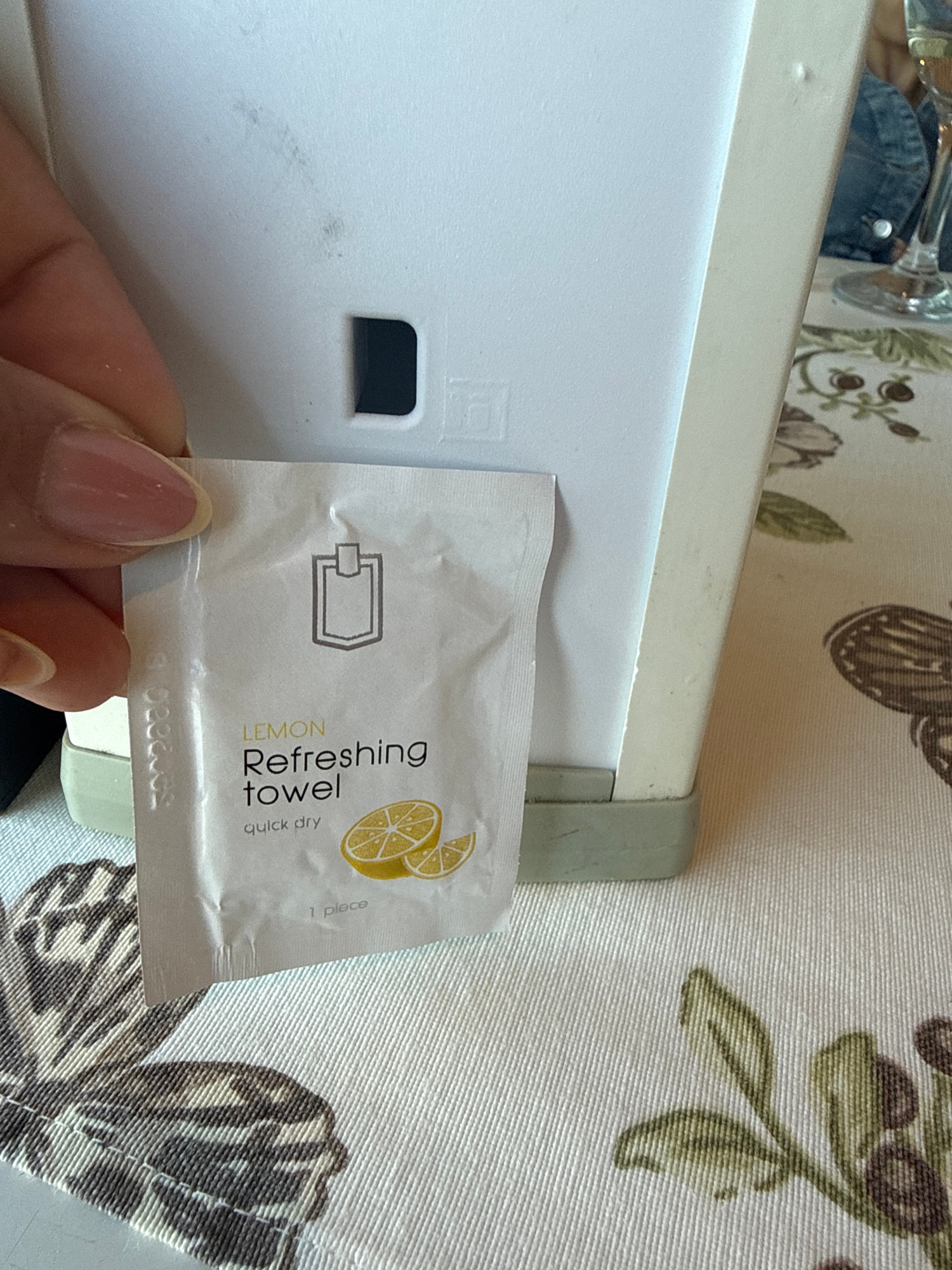 A close-up, eye-level shot shows a hand holding a single, individually wrapped refreshing towel with lemon scent. The towel packaging is white with text in black and yellow, featuring the words "LEMON Refreshing towel quick dry" and an illustration of sliced lemons. The towel is positioned in front of a white, rectangular device with a dark rectangular cutout. The device rests on a light green base. In the background, a patterned tablecloth with brown and green motifs is visible, along with a partial view of a person wearing a denim jacket and a wine glass filled with a clear liquid. The lighting is bright and diffused.