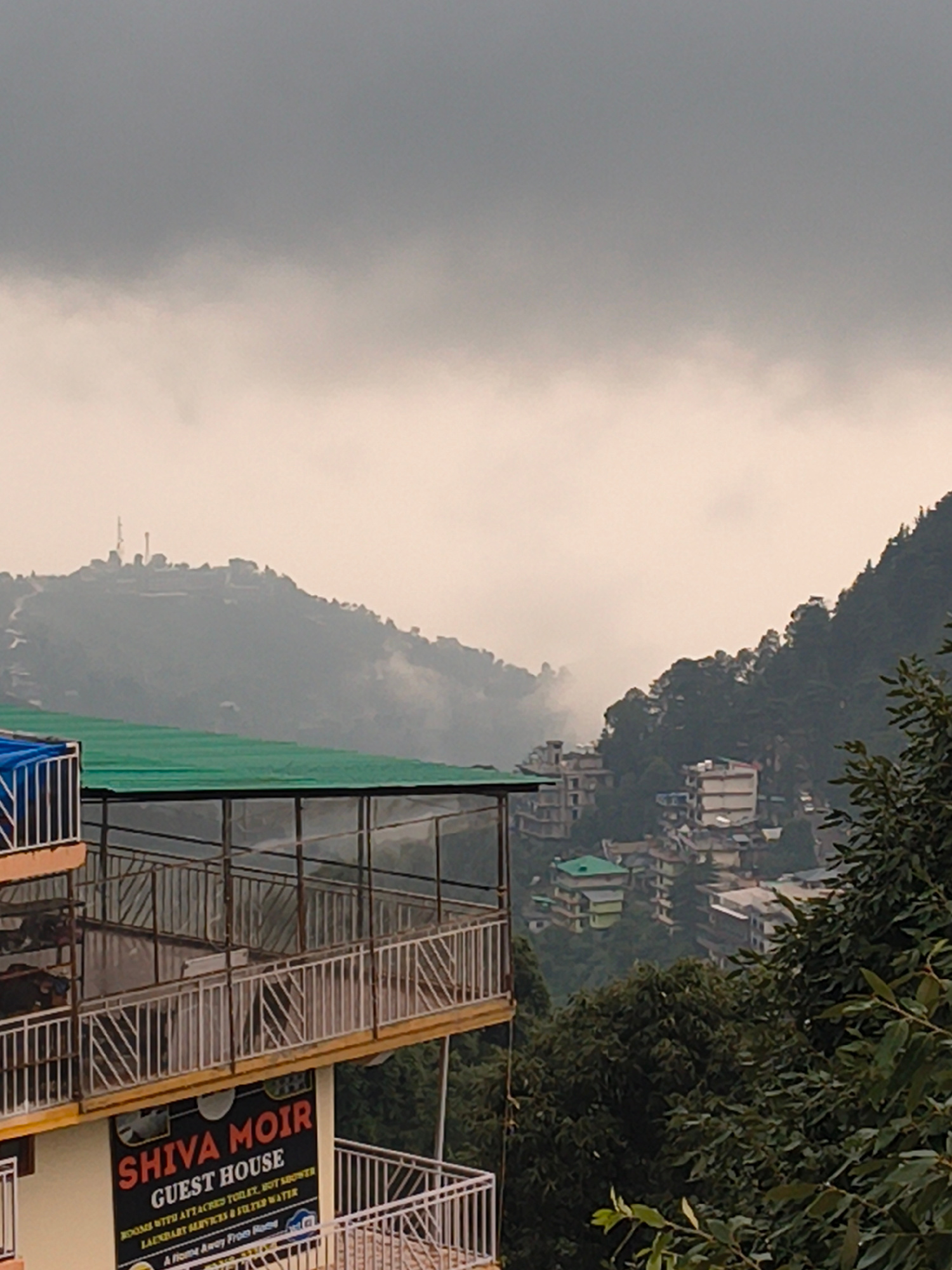 This image captures a hillside view in Dharamkot, India, under an overcast sky. The scene is dominated by a steep, tree-covered mountain slope dotted with buildings. In the foreground, a multi-story structure with white railings and a green tarp is visible, suggesting construction or renovation. A prominent sign reads "SHIVA MOIR GUEST HOUSE," further identifying the location. The weather appears to be cloudy, casting a soft light over the landscape. The presence of the guest house indicates it is likely a tourist or residential area. The overall mood is serene, with the soft light and the natural scenery creating a peaceful atmosphere. The image is vertically oriented, emphasizing the height of the mountain and the layout of the buildings.