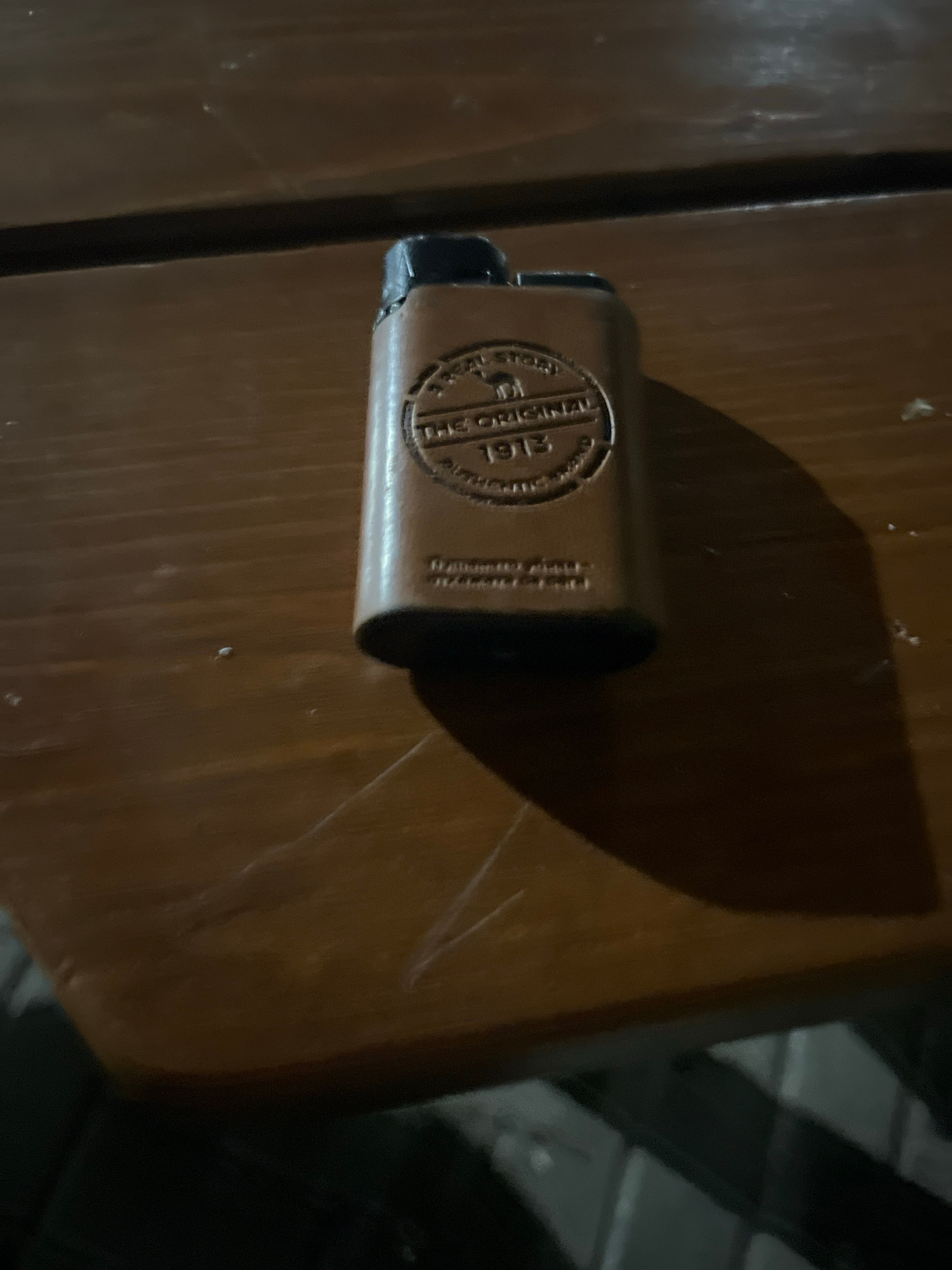 A light brown, rectangular object, possibly a lighter case or a small pouch, with embossed lettering and a circular emblem featuring a camel, rests on a dark brown wooden surface. The lettering around the emblem reads "THE ORIGINAL 1913" and appears to have additional text below it. The object is positioned diagonally on the surface, with the top part closer to the viewer and casting a shadow. The background is out of focus and appears to be a dark, textured surface, possibly a table or floor, with some faint lighting. The image was captured in Bulgarovo, Bulgaria.