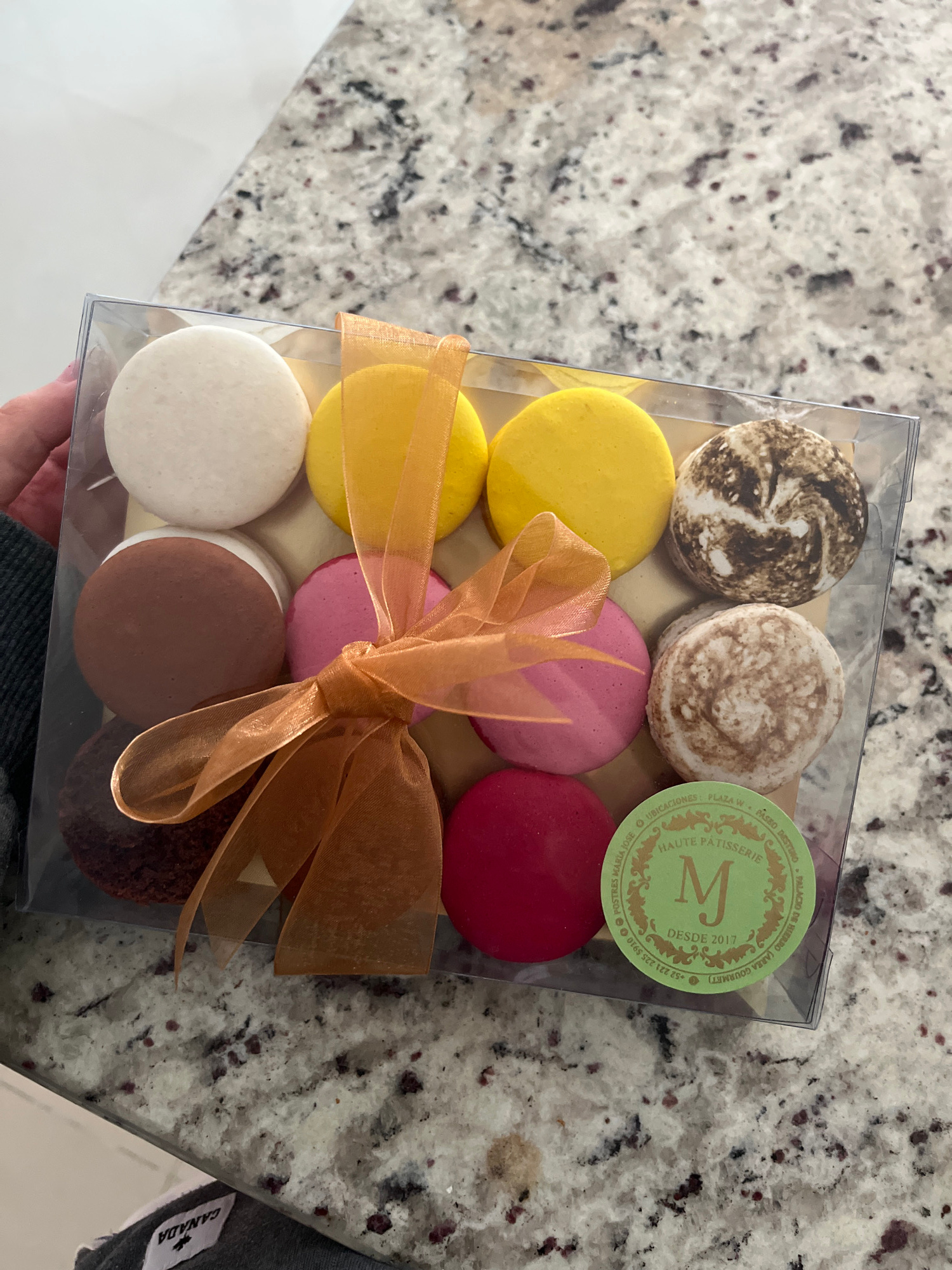 A clear plastic box containing a dozen assorted macarons is presented. The macarons are arranged in two rows of six, with the top row featuring a white macaron, two yellow macarons, and two speckled brown and white macarons. The bottom row displays a brown macaron, a pink macaron, a reddish-pink macaron, and a deep red macaron. A bright orange ribbon is tied in a bow around the box, partially obscuring some of the macarons. A circular green sticker with gold lettering is affixed to the lower right corner of the box, displaying the text "HAUTE PATISSERIE" and "DESDE 2017" along with the initial "M" in ornate gold lettering. The box rests on a speckled granite countertop, the texture of which is characterized by a gray base with dark brown and black flecks. In the bottom left corner, a portion of a gray garment with a white label featuring a maple leaf symbol and the word "CANADA" is visible. The overall scene is lit from above, casting soft shadows. The location is identified as Barrio de Nuevo León, Mexico, specifically referring to the "Postres Maria Jose" establishment where the macarons are sold, as indicated by the sticker.