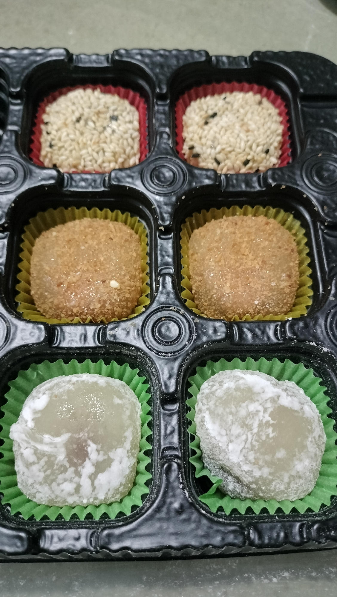 A black plastic tray holds six mochi. The tray is divided into six individual compartments. The top row contains two mochi covered in sesame seeds, one with black sesame seeds, and the other with white and black sesame seeds. These are nestled in red paper liners. The middle row has two mochi covered in a sandy brown coating, likely crushed nuts or cookie crumbs, placed in yellow paper liners. The bottom row contains two pale, white mochi that appear dusted with a white powder, possibly cornstarch or confectioners' sugar, in green paper liners. The mochi are arranged in a 2x3 grid. The background is a neutral, grey surface, suggesting the tray is on a table or countertop. The lighting appears to be indoor and even. There is no text visible in the image. The scene depicts a collection of sweet treats, possibly for consumption or as part of a dessert offering. The location context of Medan, Indonesia, does not provide specific visual cues within the image itself.