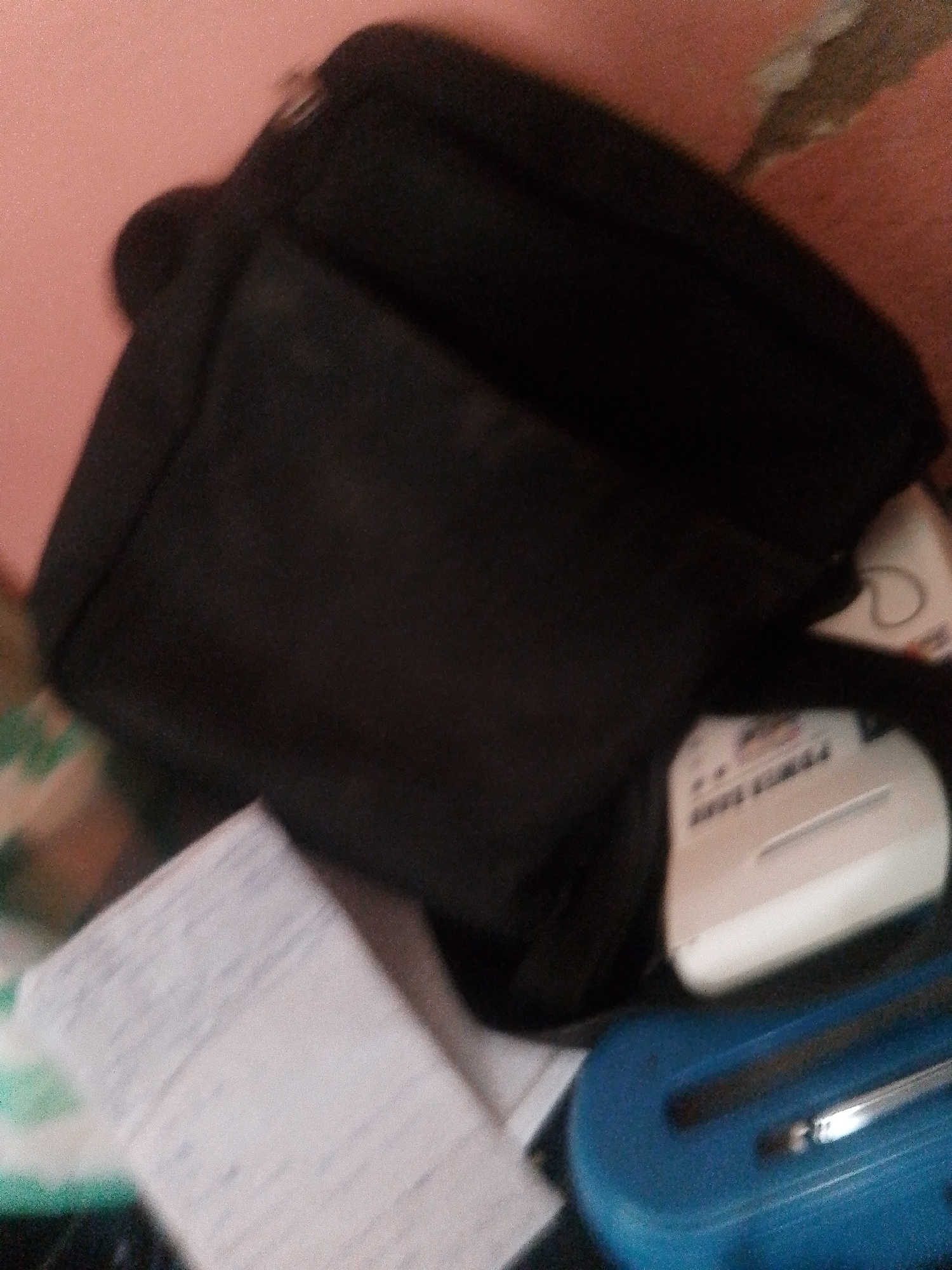 The image shows a close-up, slightly blurry view of a black bag, likely a messenger bag or a satchel, resting on a surface. To the right of the bag, a white object with some text and a metallic slot is visible, possibly a small shredder or a similar electronic device. Below the bag, a stack of papers with handwritten notes is also present. The background is a pinkish-orange wall. The overall impression is of a personal workspace or a collection of items belonging to an individual. No specific activity is discernible from the image. The lighting appears to be artificial, suggesting an indoor setting. There is no clear indication of the time of day or weather conditions. The location context of Jauro Yinu, Nigeria, is provided, but no specific visual cues within the image directly confirm this location. The visible text on the white object is partially obscured and illegible.