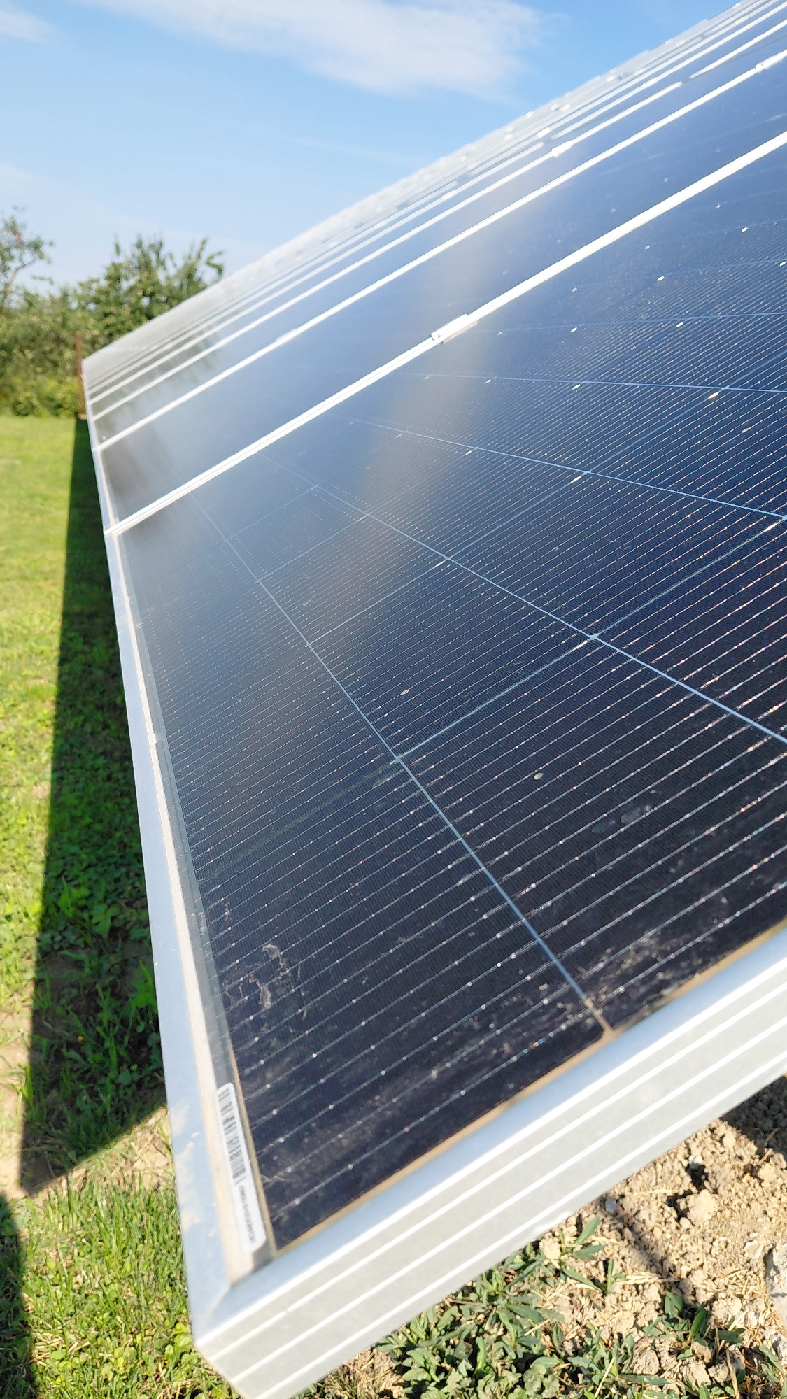 The image shows a close-up, low-angle view of a solar panel array under a clear blue sky with scattered clouds. The solar panels are dark blue with a grid pattern of thin wires. They are mounted on a silver-colored frame, which is supported by the ground. The foreground consists of green grass and dry soil, with some small weeds growing. In the background, out of focus, are green trees and a shadow cast by the solar panels. The scene appears to be outdoors in a rural or semi-rural setting. There are no people or animals visible in the image. The image is taken in Uzhhorod, Ukraine.