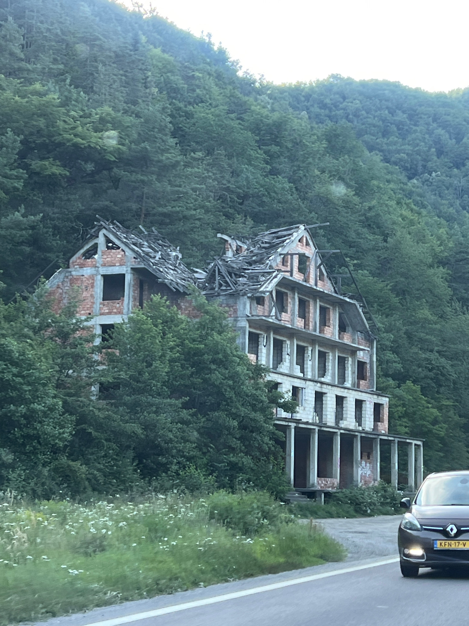 The image shows an abandoned, multi-story building with a damaged roof, set against a backdrop of lush green trees on a hillside. The building appears to be constructed of brick and concrete, with visible structural damage and incomplete construction. The windows are empty openings. In the foreground, a dark-colored car with a license plate reading "KFN-17-V" is driving on a road. A field with white wildflowers is visible between the road and the building. The location is identified as Copăceni, Romania.