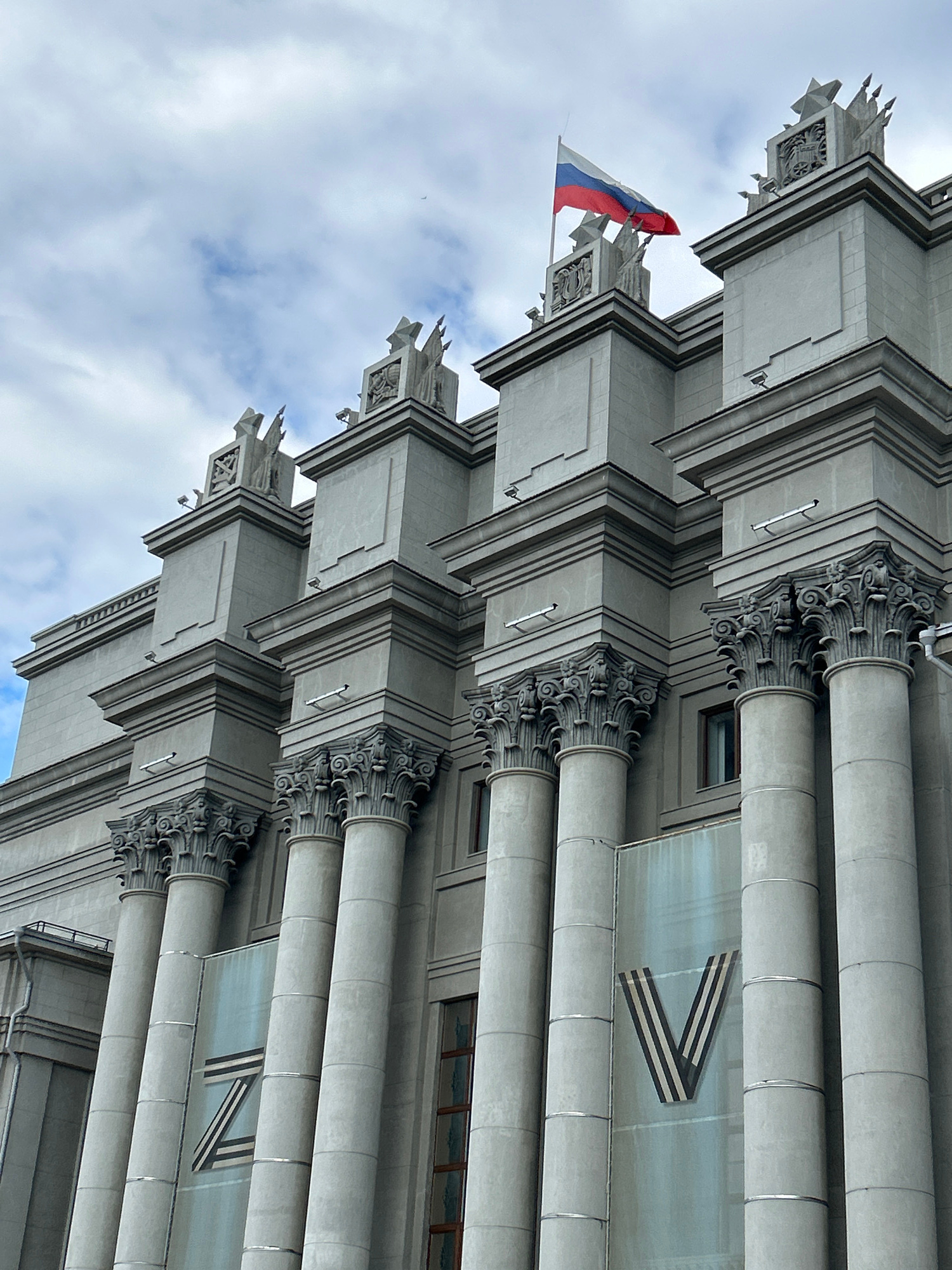 The image shows the facade of the Novokuybyshevsk Palace of Culture, a grand building with a classical architectural style. The building features large, fluted columns topped with ornate Corinthian capitals. Above the columns, there are decorative sculptures and a Russian flag waving from a flagpole. Two large stylized letters, "Z" and "V", are visible on banners or glass panels attached to the building's facade between the columns. The sky above is cloudy, with patches of blue. The overall impression is of a solemn and imposing structure.