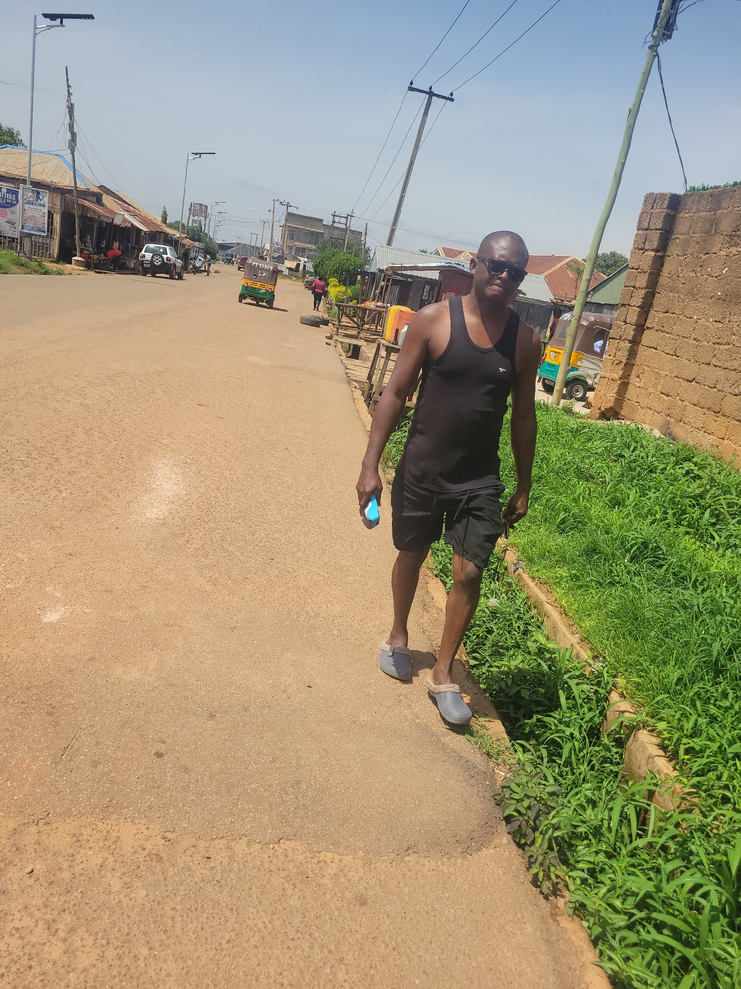 A man wearing a black tank top and black shorts walks on the side of a dusty, paved road in Jos, Nigeria. He is also wearing gray slippers and sunglasses. He holds a light blue object in his right hand. To his left, the road continues, with shops lining the side. A white SUV and a few other vehicles are visible in the distance. To his right, there is a patch of tall green grass and a brick wall. The sky is clear and blue, and there are power lines and utility poles visible throughout the scene. A yellow and green tuk-tuk is driving down the road in the background.