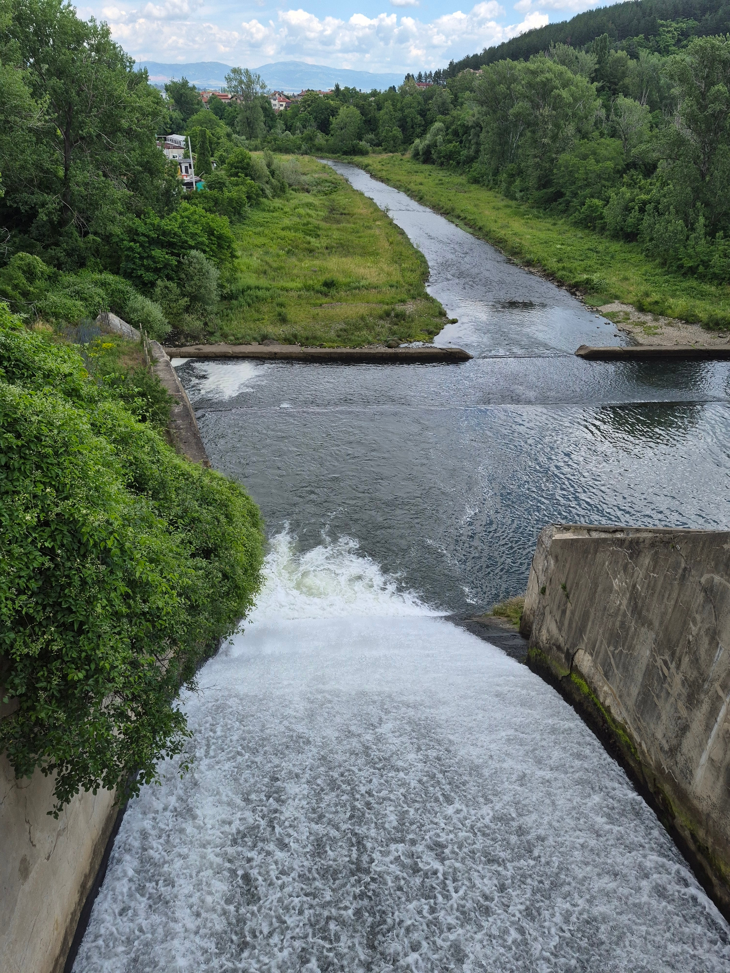 A high-angle, vertical, full shot captures a wide, powerful waterfall cascading down a concrete dam. The turbulent white water fills the foreground, frothing and churning as it descends. On the left, lush green foliage spills over the edge of the dam, contrasting with the gray concrete structure. The dam itself is reinforced with concrete walls, showing signs of age with cracks and mossy patches.
Beyond the dam, a river flows into the distance, its surface rippling. Further upstream, the river narrows and meanders through a verdant landscape of grassy banks and dense trees. The river appears to be contained within a channel with concrete embankments in places. In the far background, a town with red-tiled roofs is nestled amongst hills covered in trees, and distant mountains form the horizon under a sky with scattered white clouds. The scene is a natural landscape enhanced by man-made structures, a dam controlling the flow of a river within a semi-rural or suburban environment. There are no people or animals visible in the frame.